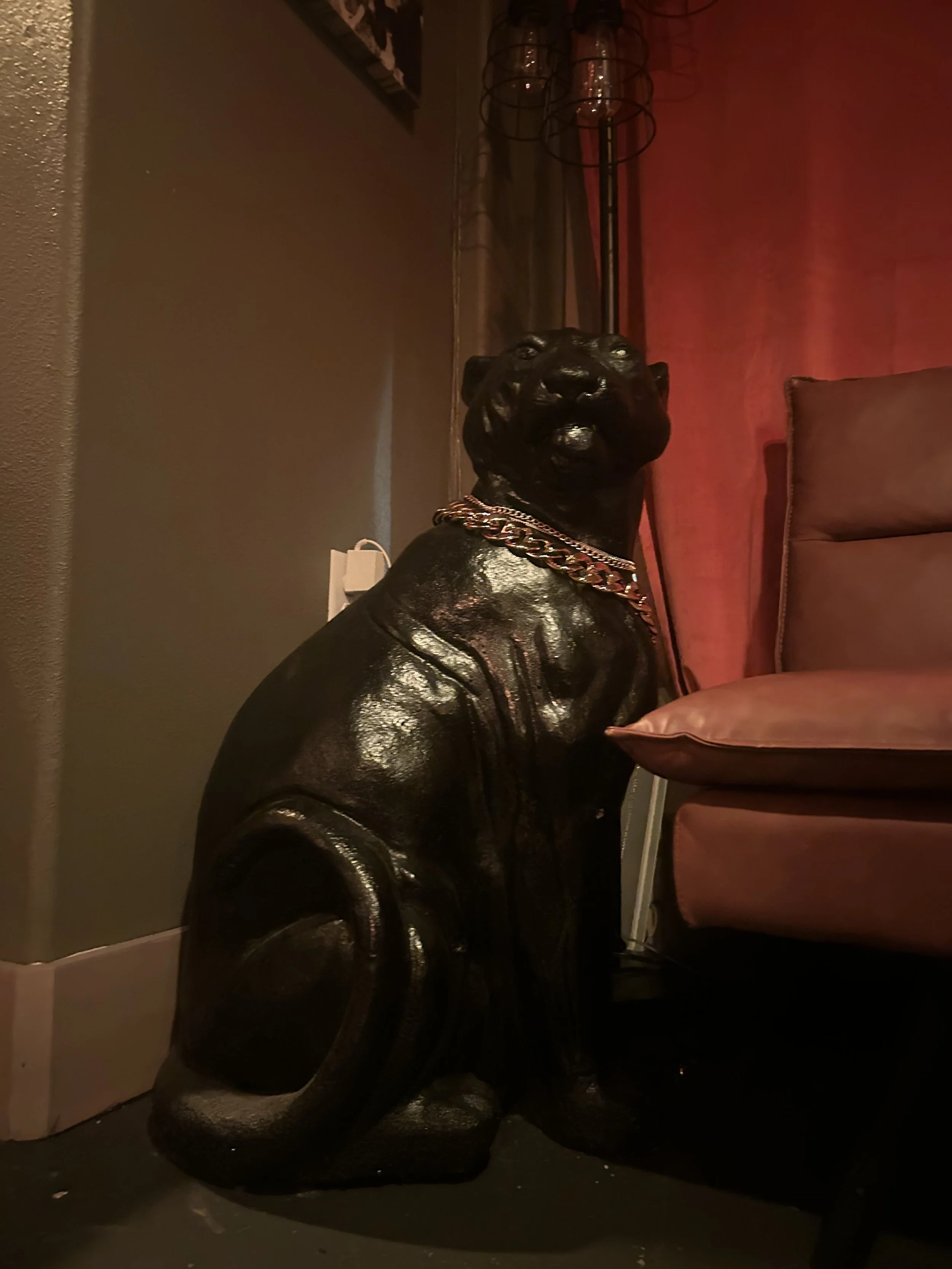 A black statue of a sitting dog, possibly a panther or a similar large cat, wearing a gold chain necklace, positioned next to a pinkish armchair in a dimly-lit room with dark and warm tones.
