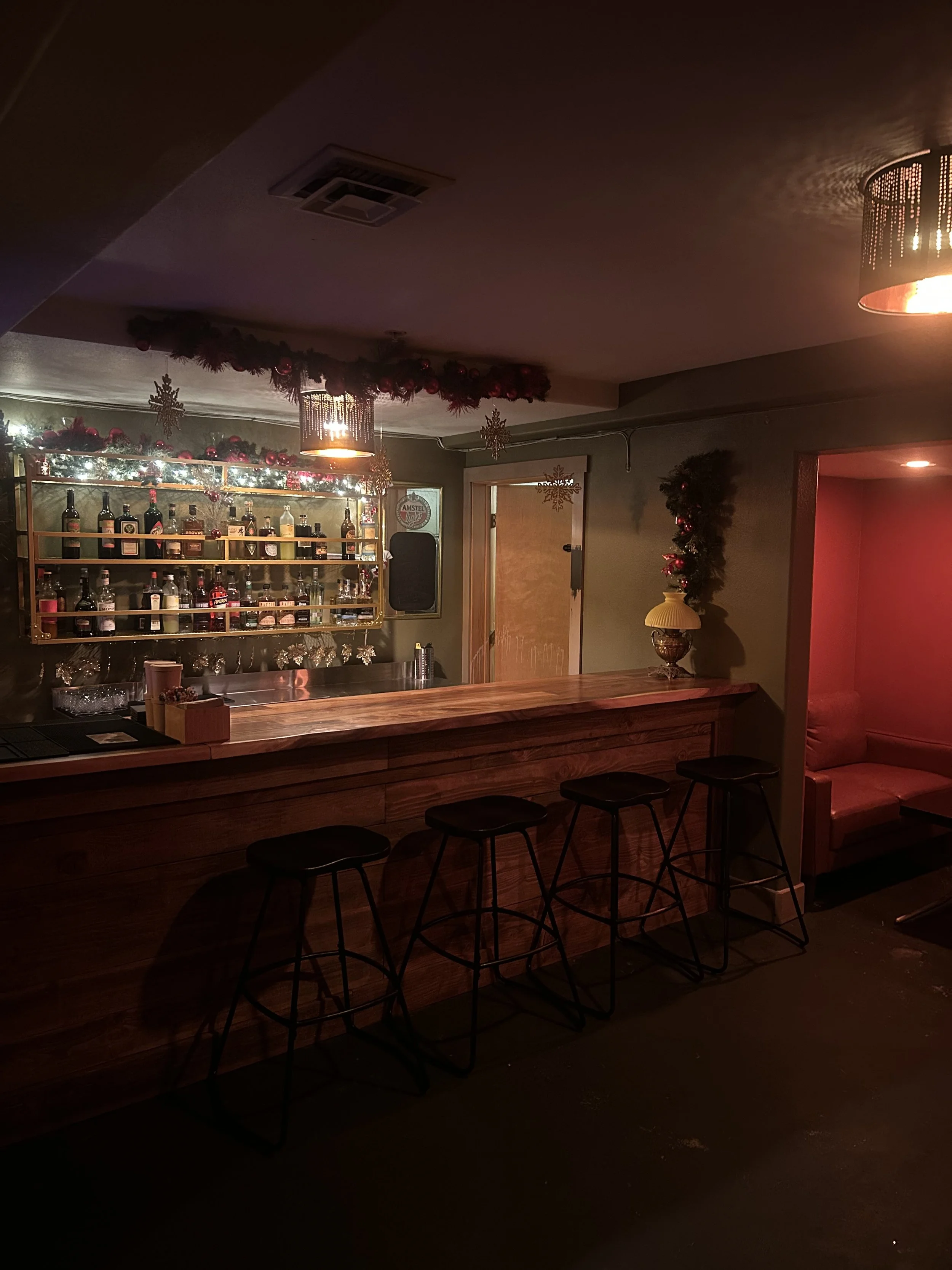Empty bar area decorated with Christmas ornaments, including garlands, snowflakes, and lights, with a wooden counter and four bar stools.