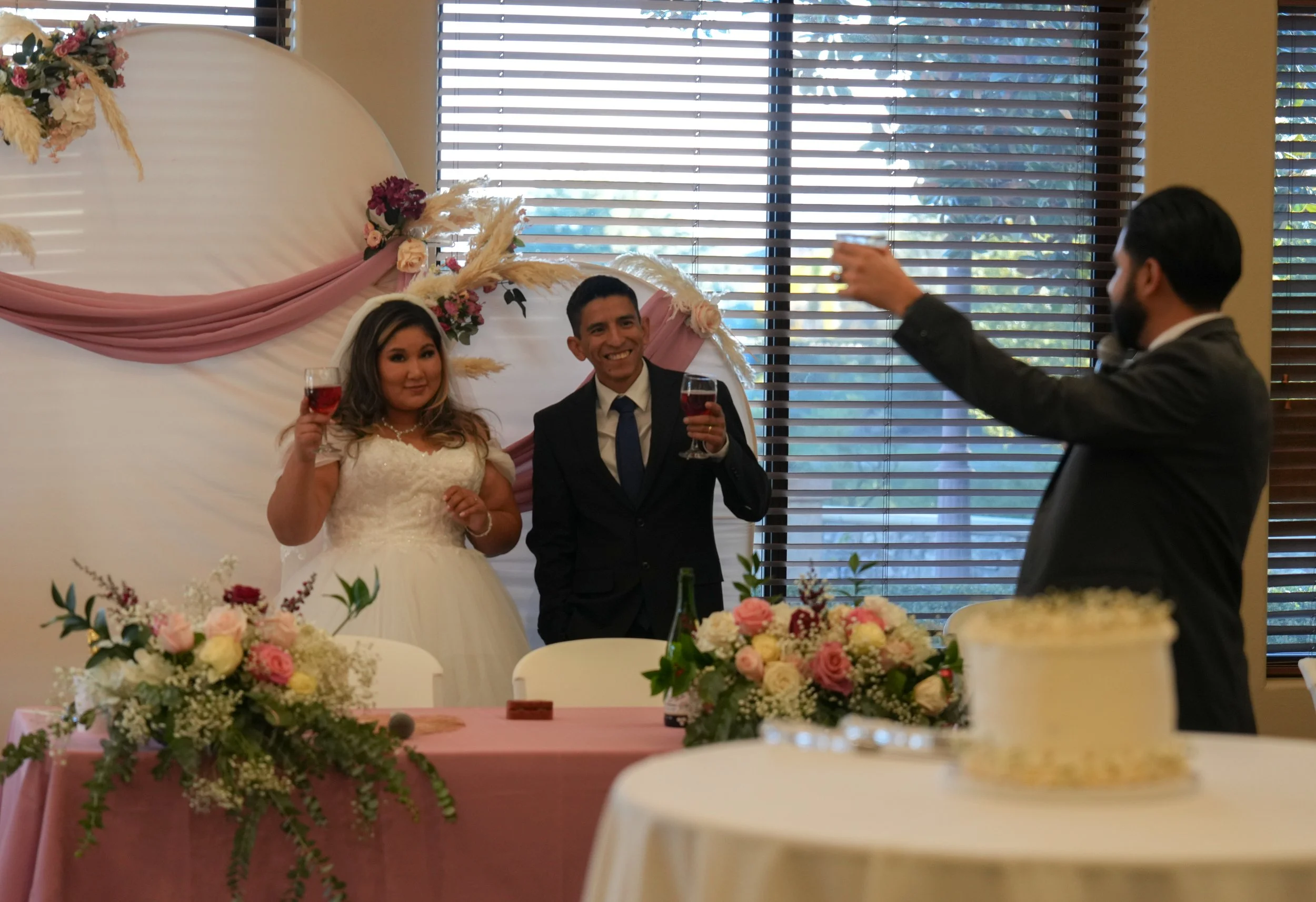 Bride and groom raising glasses in toast at wedding reception, sitting at decorated table with floral arrangements, behind a backdrop with draped fabric and floral accents