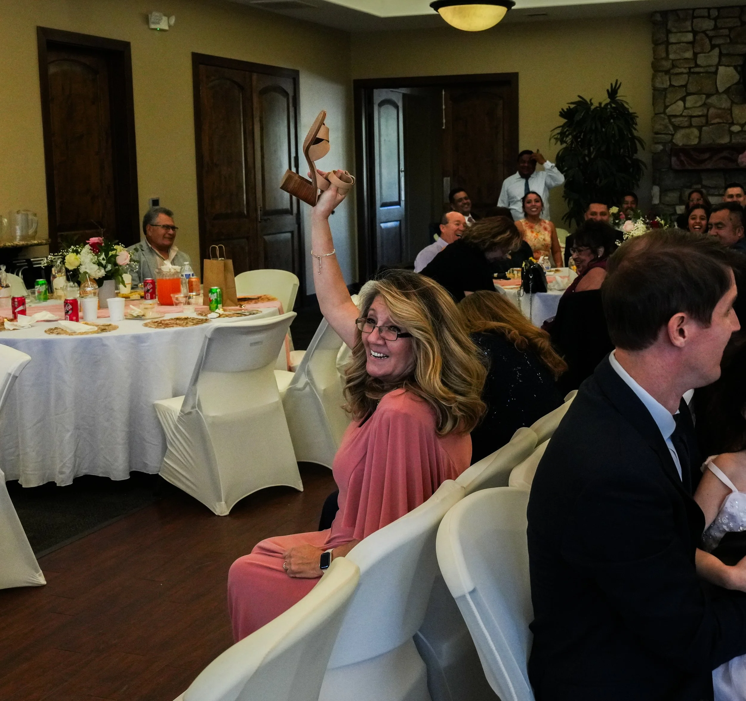 A woman in a pink dress sitting at a banquet table raising her heel in a celebration, with guests laughing and smiling in the background at a formal event or wedding reception.