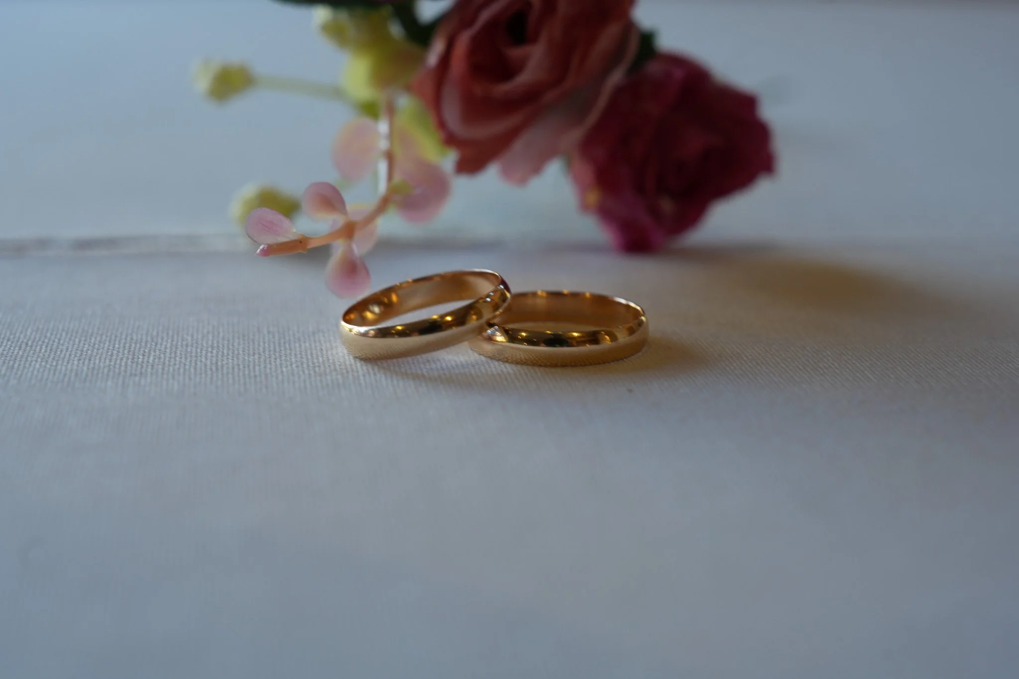 Two gold wedding rings placed on white fabric with pink and red flowers in the background.