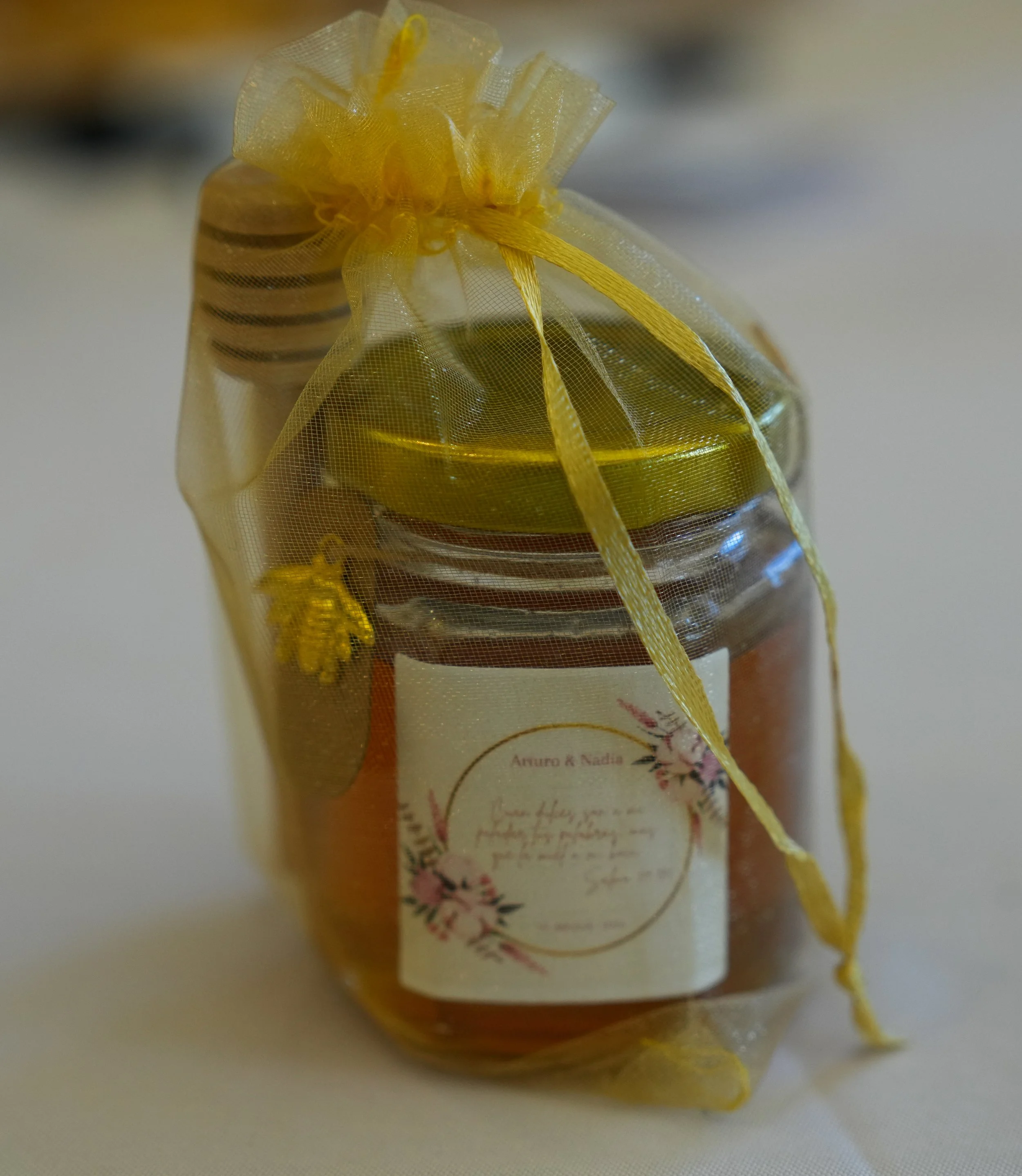 A small jar filled with honey, wrapped in a yellow organza gift bag with a bow, and a gold honey dipper on top.