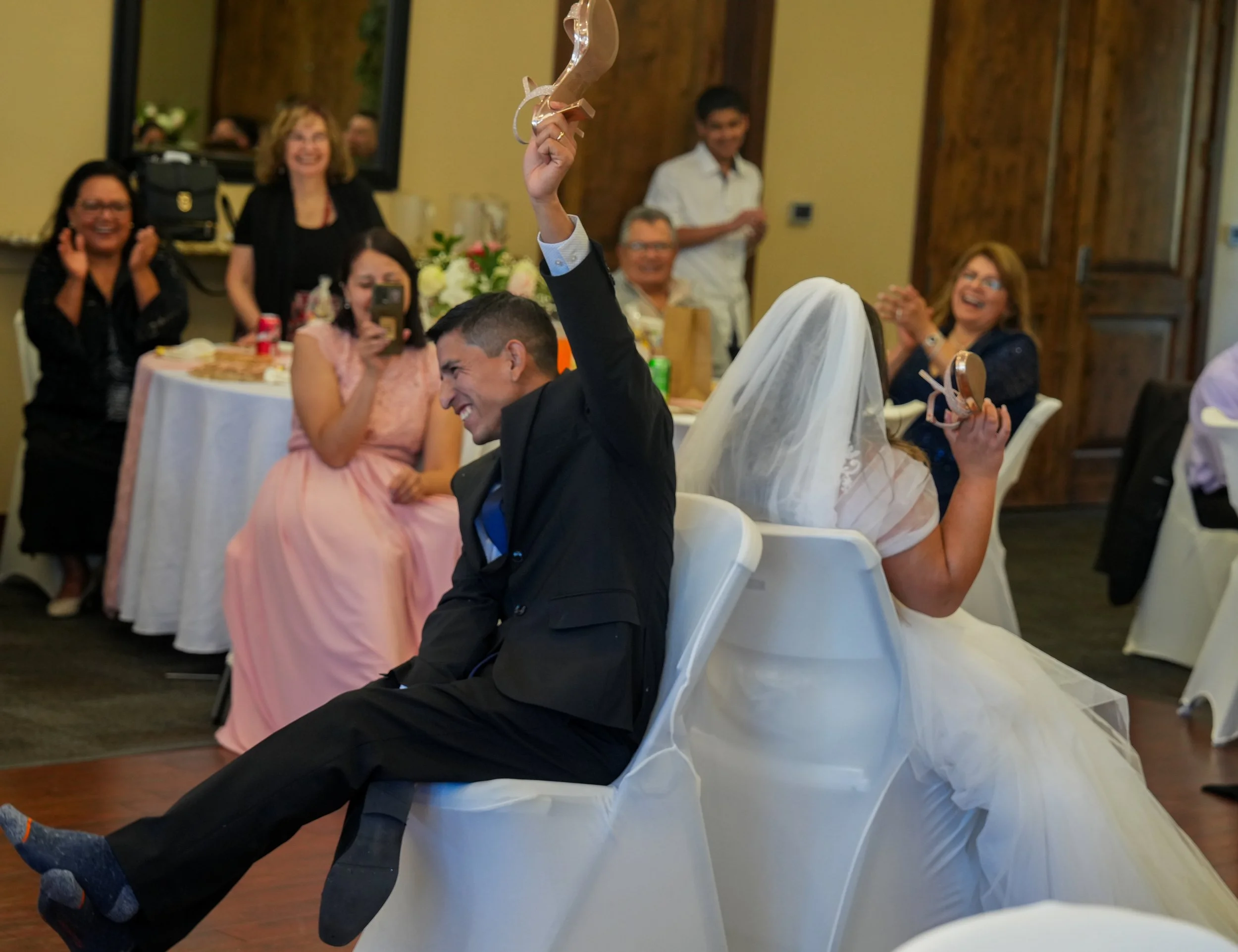 A bride and groom sitting back-to-back in chairs at a wedding reception, playing a game with small hats or headbands on sticks. Guests are seated around tables, smiling and laughing, capturing the moment with their phones.