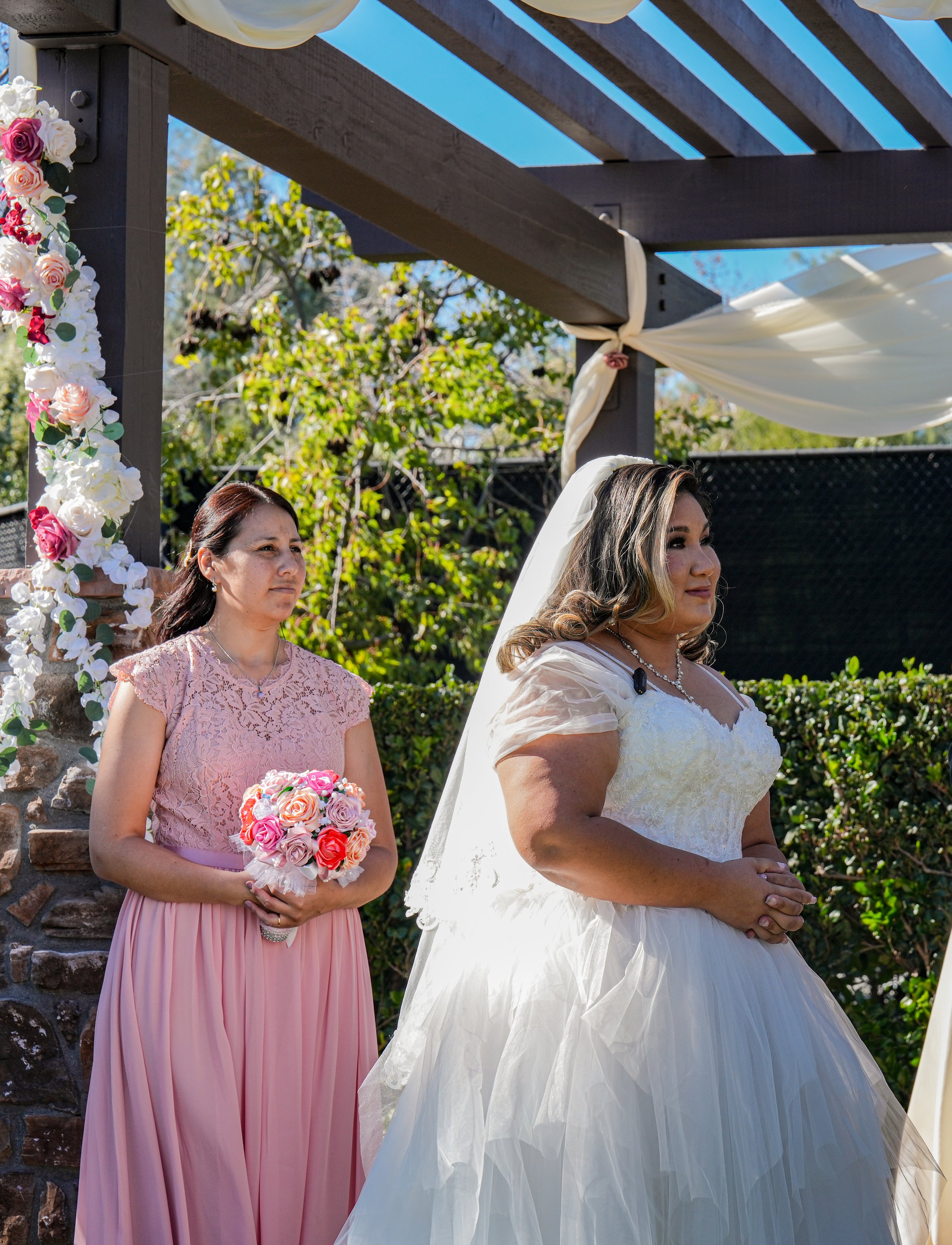 A bride in a white wedding gown, wearing a veil, stands outdoors with her hands clasped. Beside her is a woman in a pink lace dress holding a bouquet of pink, red, and white roses. They are under a pergola decorated with white fabric and flowers, wit