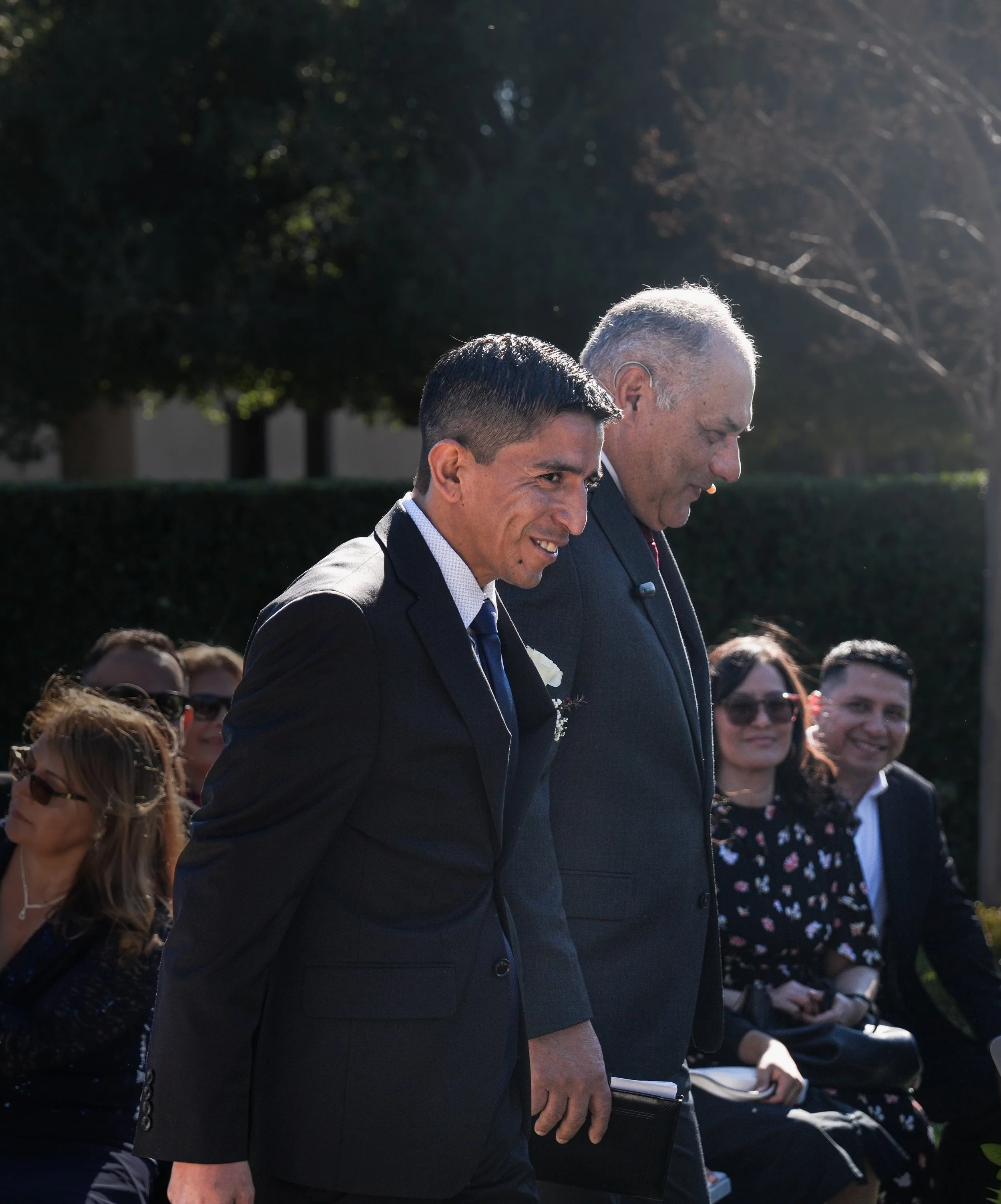 Two men in suits walking outdoors during a formal event, with seated attendees in the background.