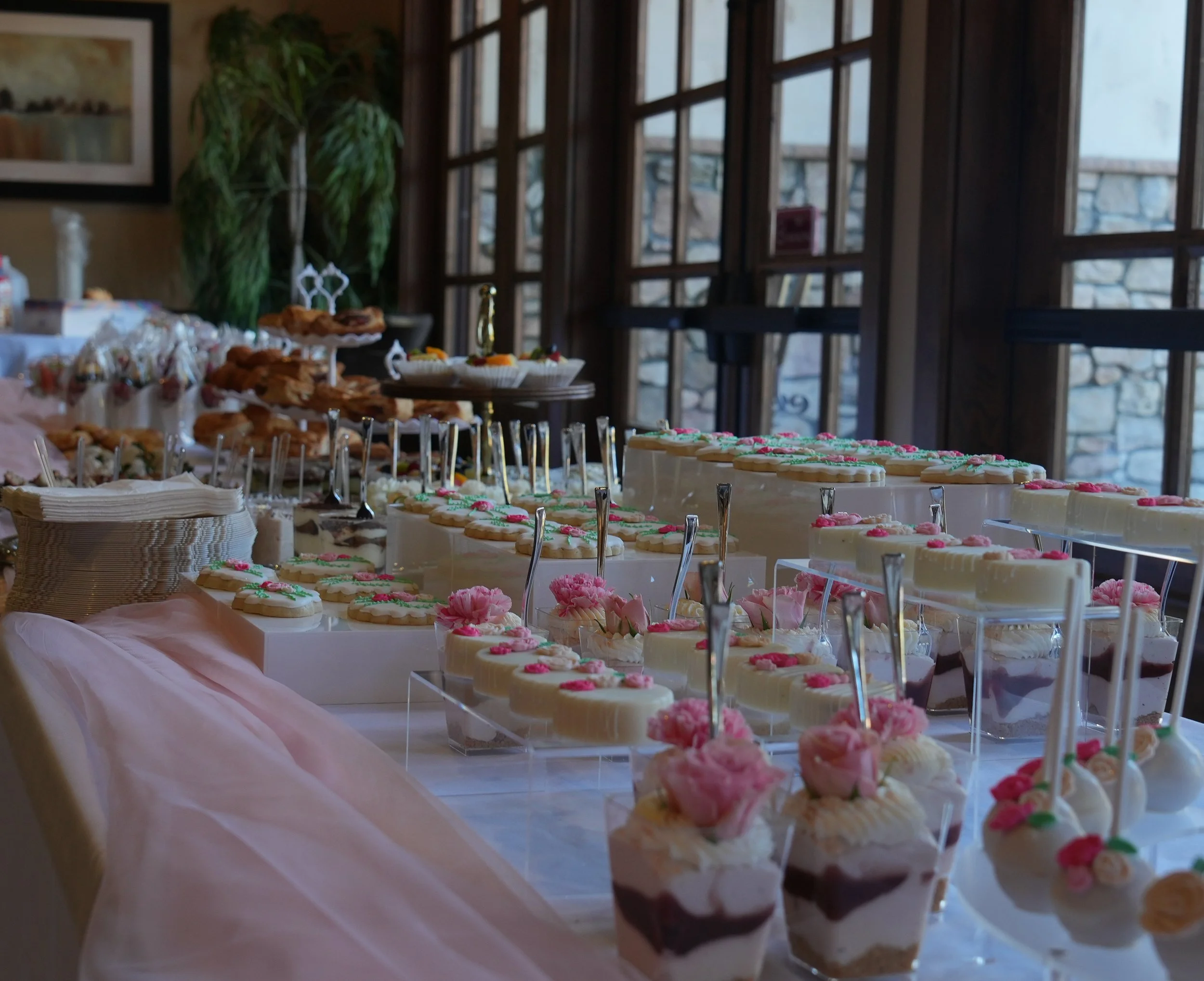A dessert table with various decorated sweets, including cupcakes, cake pops, and layered cakes, set in a room with large windows and natural light.