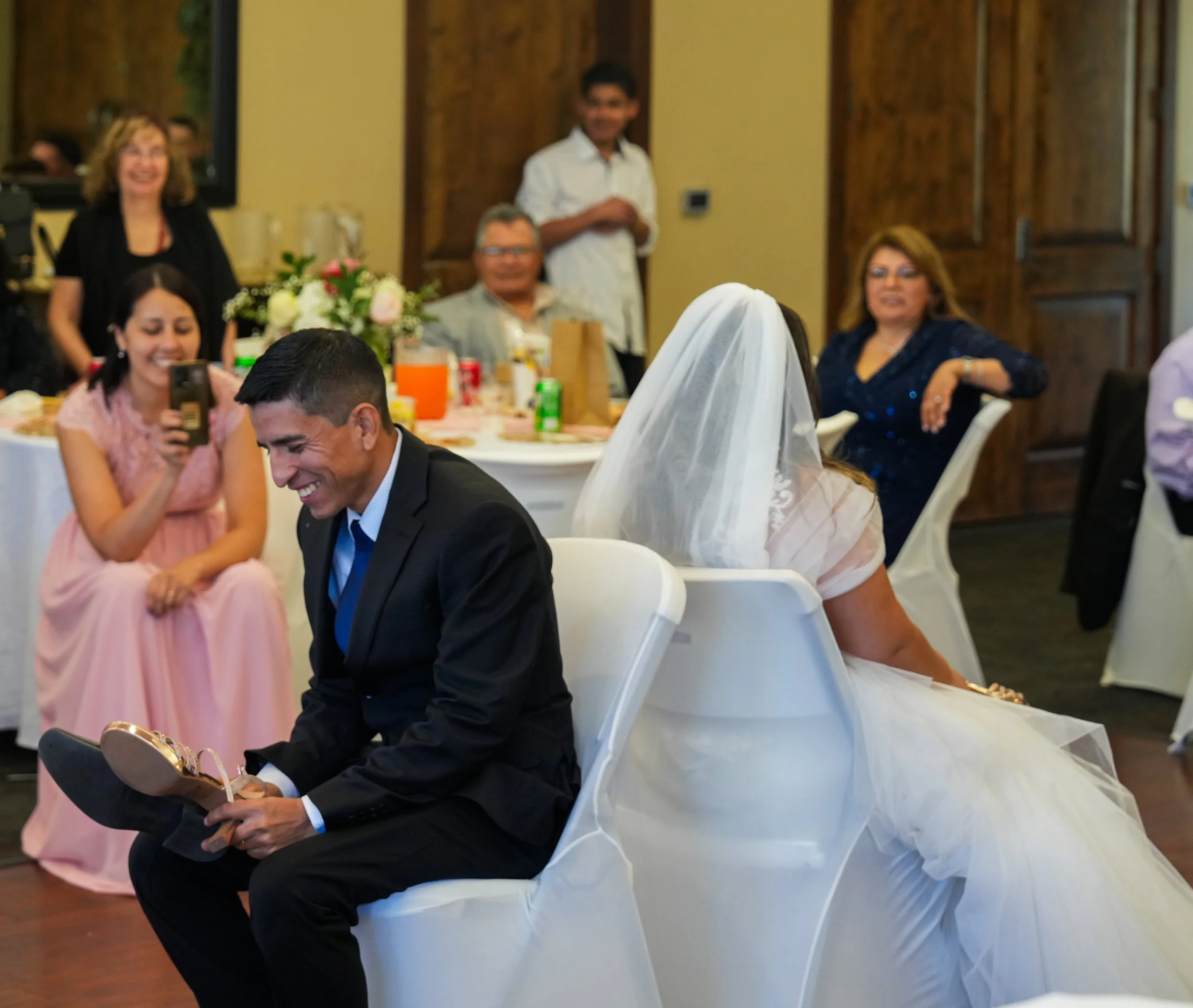 A bride in a wedding dress and veil seated next to a man in a suit, both smiling, with wedding guests in the background at a reception.