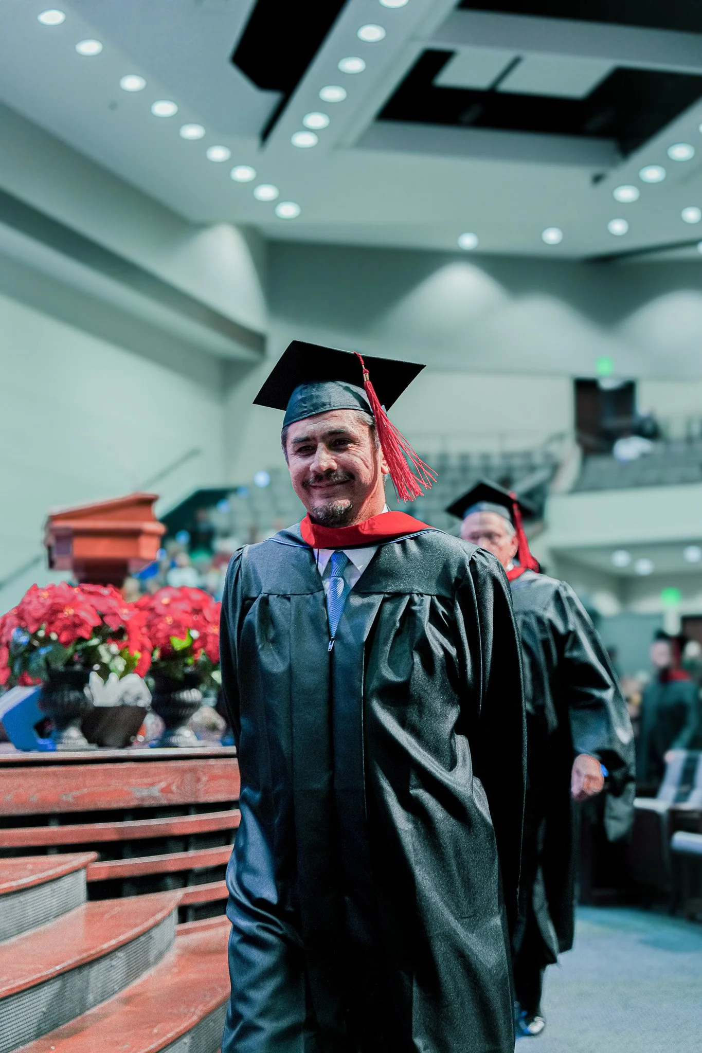 A man in a cap and gown at a graduation ceremony, smiling, with other graduates in gowns and caps in the background, indoors with bright lighting and seating.
