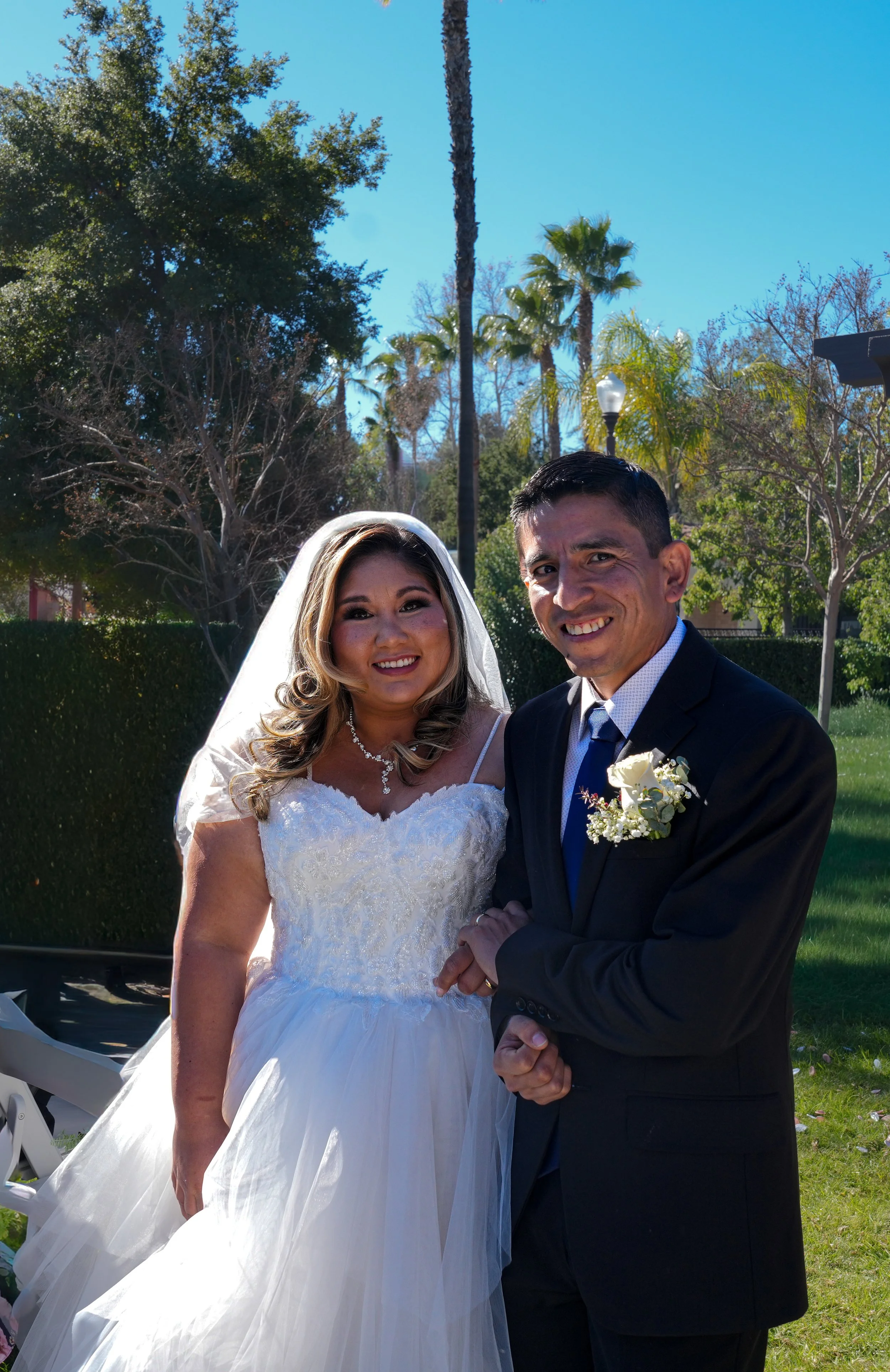A bride and groom smiling for a photo outdoors on a sunny day, with trees and palm trees in the background.