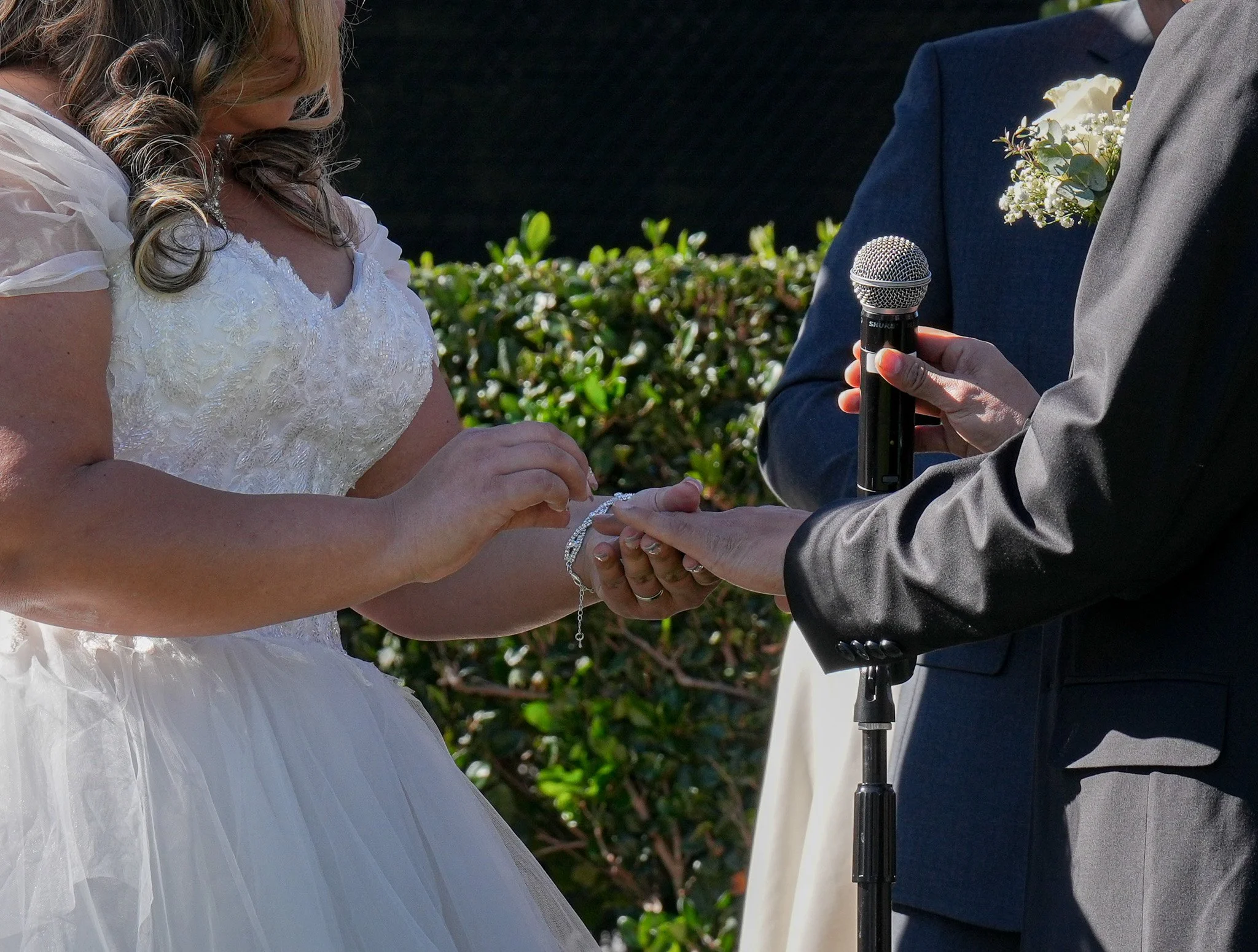 Bride and groom exchanging rings during outdoor wedding ceremony, with officiant holding a microphone.