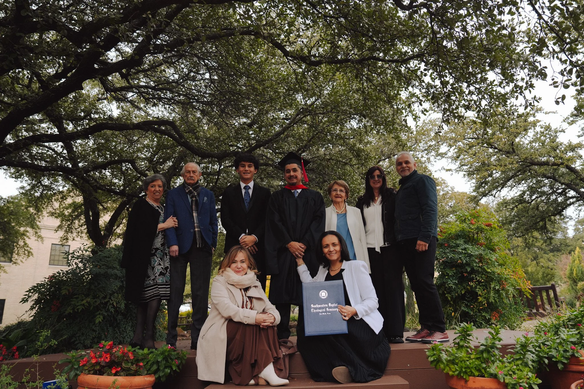 Group of nine people, including a young man in a graduation cap and gown, posed outdoors under large trees. Some are standing, and two women are kneeling in front, one holding a diploma from Southwestern Baptist Theological Seminary.