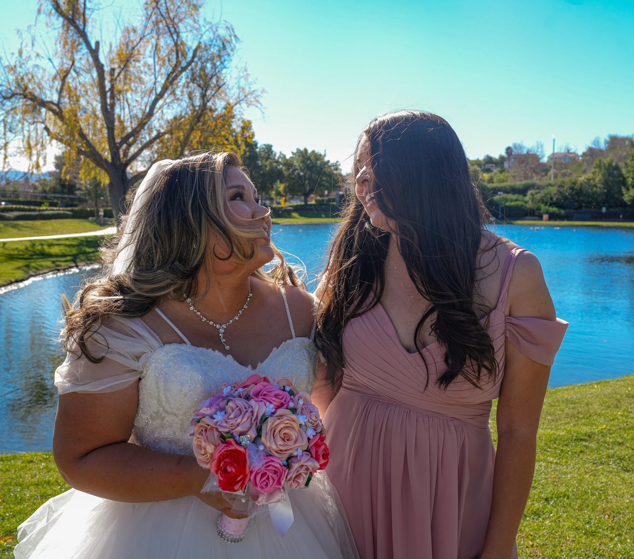 A bride in a white wedding dress holding a pink and purple bouquet, standing outdoors with a woman in a pink dress, near a pond with trees and greenery in the background.