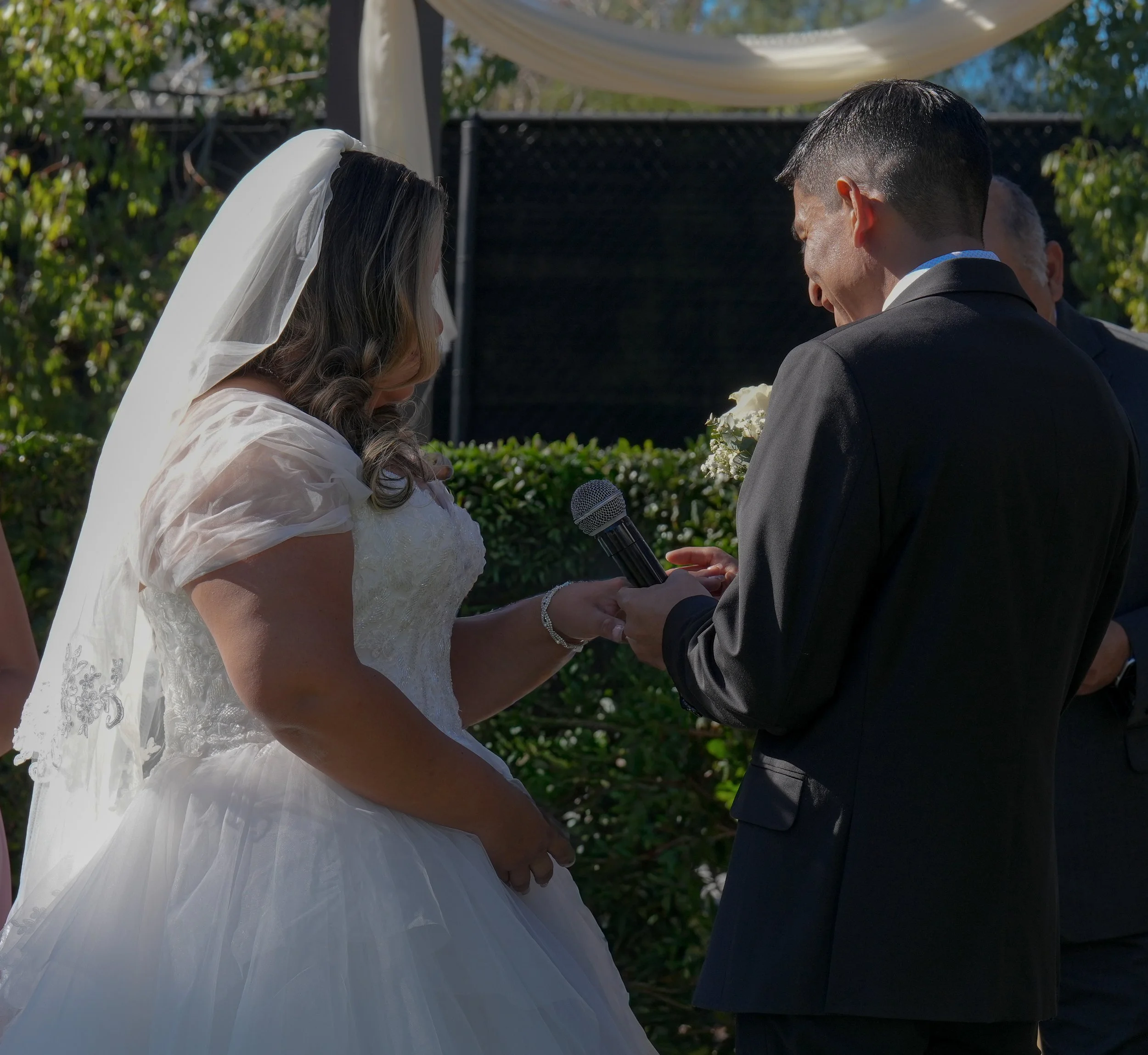 A bride and groom during their wedding ceremony outdoors, with the bride holding a microphone, and the groom holding her hand, standing close together.