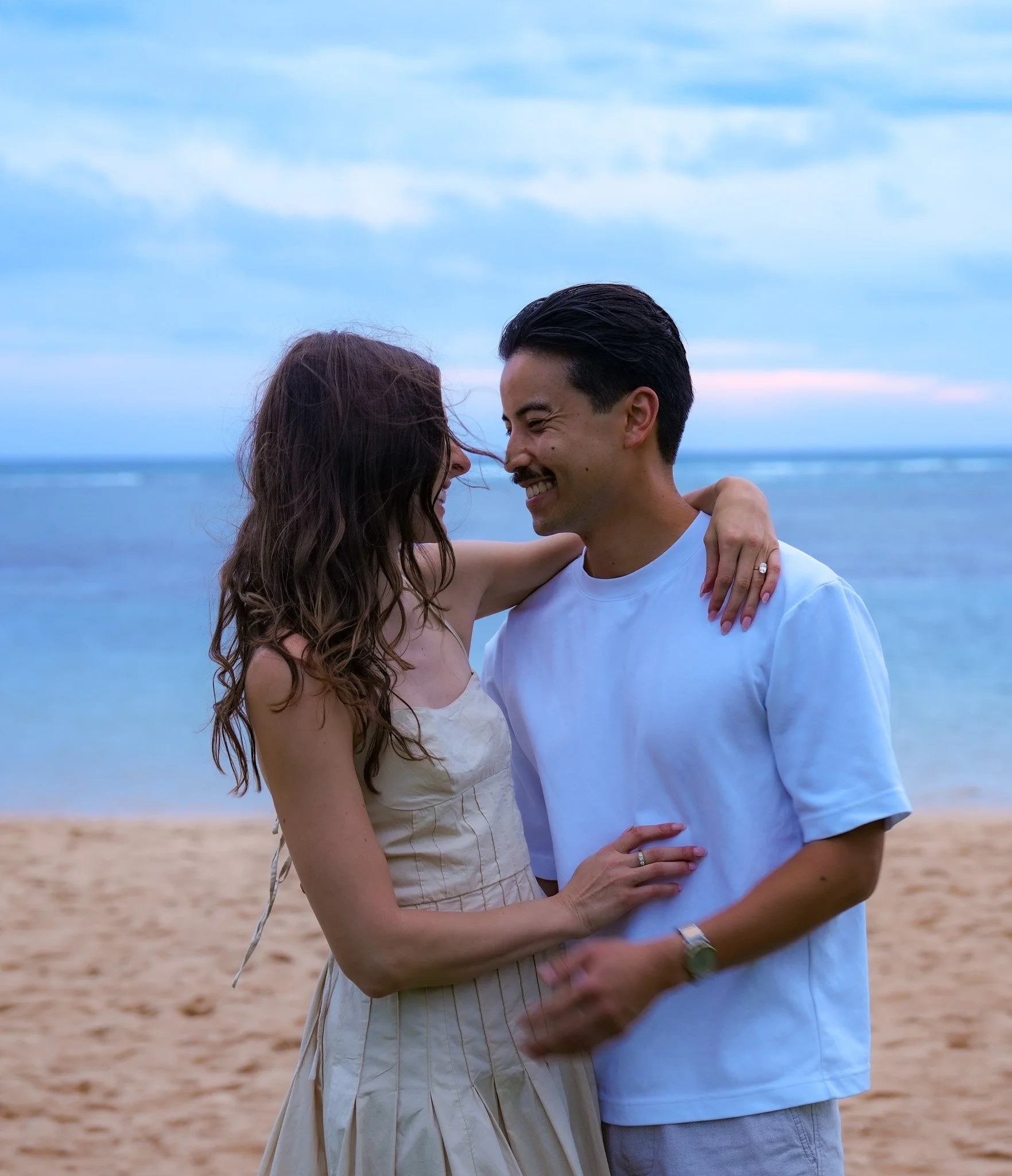 A couple smiling and looking at each other on the beach, with the ocean and sky in the background.