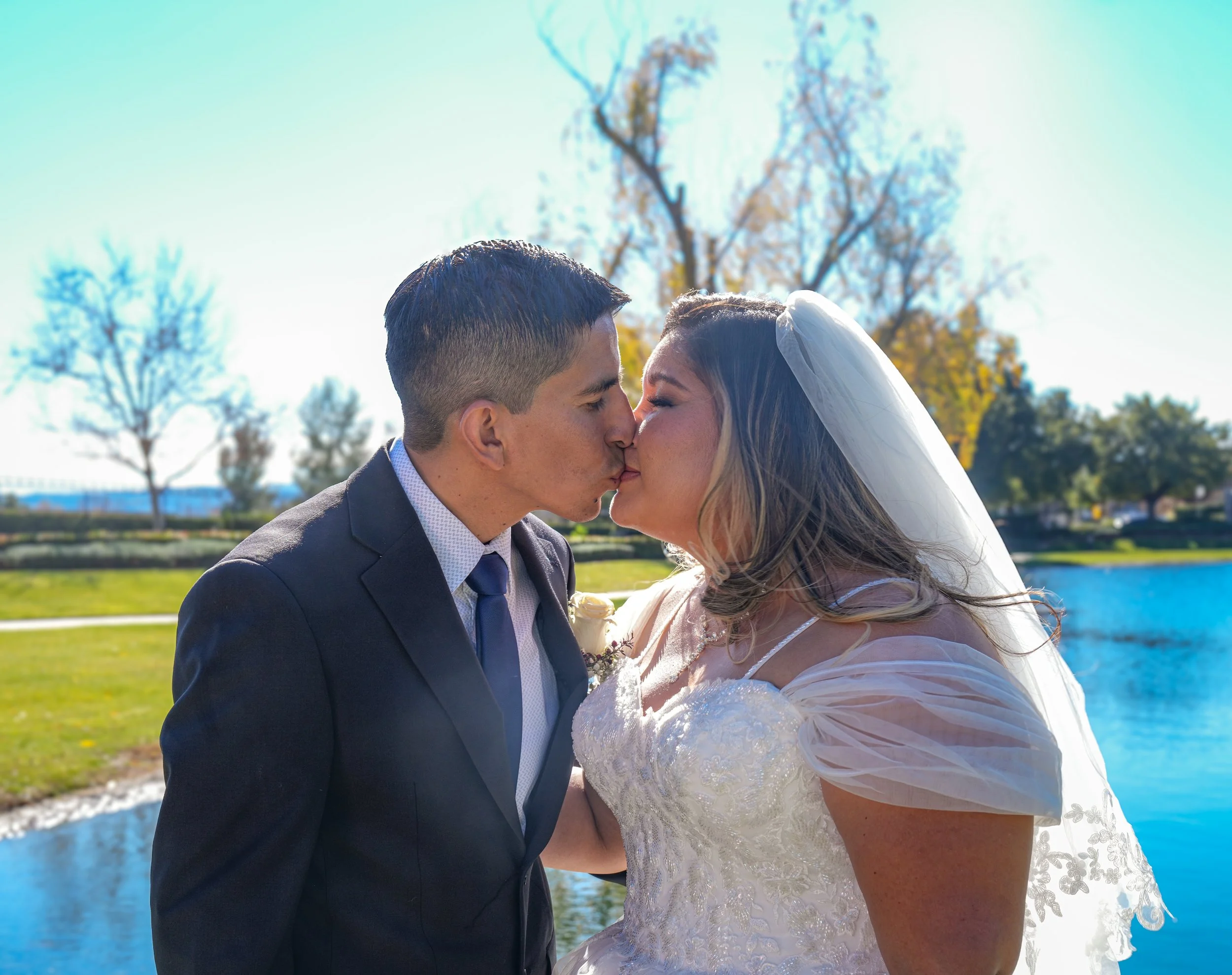 A newlywed couple kissing outdoors near a body of water with trees in the background on a sunny day.