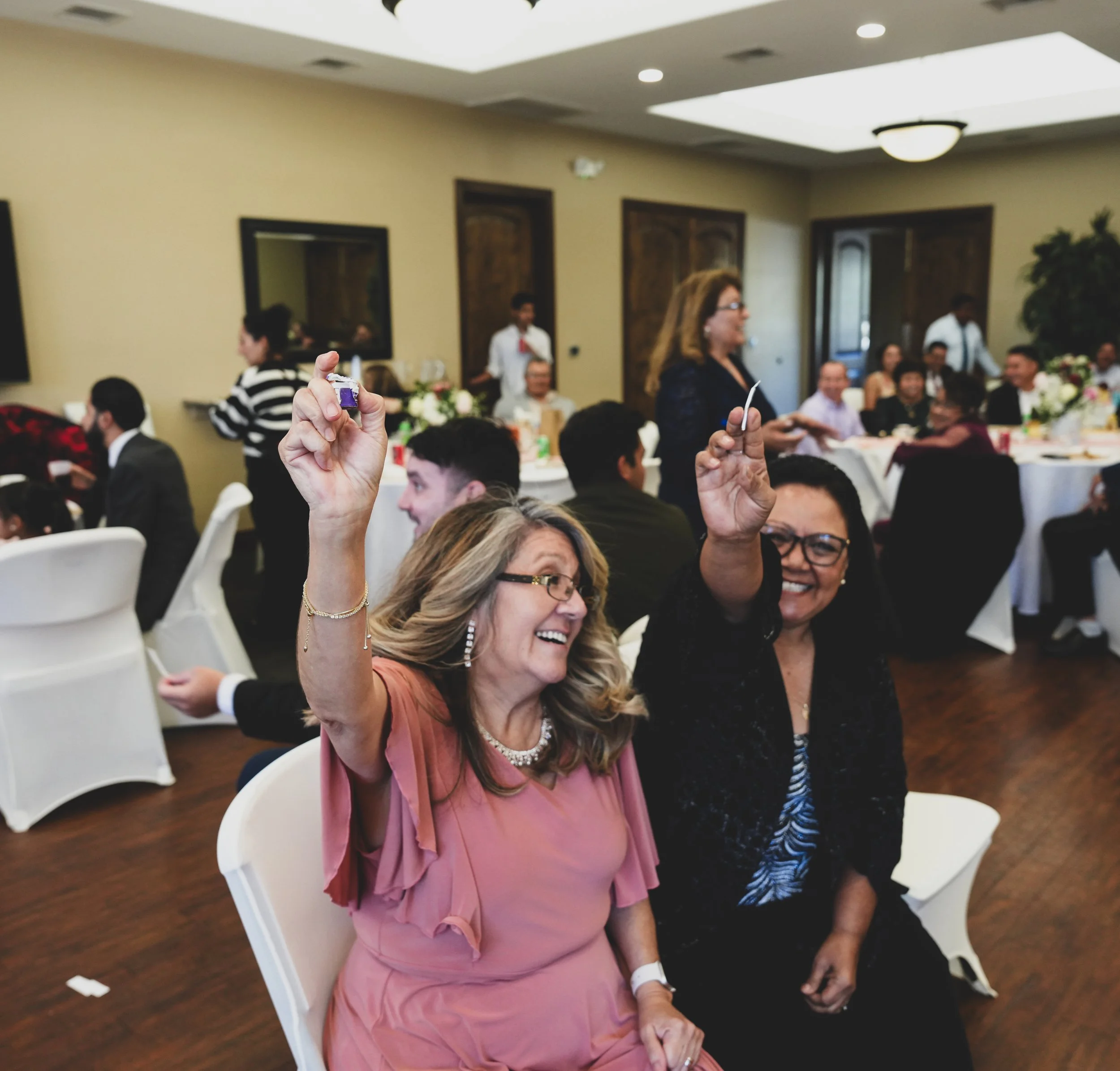 Two women celebrating at a formal event, smiling, holding up a small object, seated at a decorated table with guests in the background.