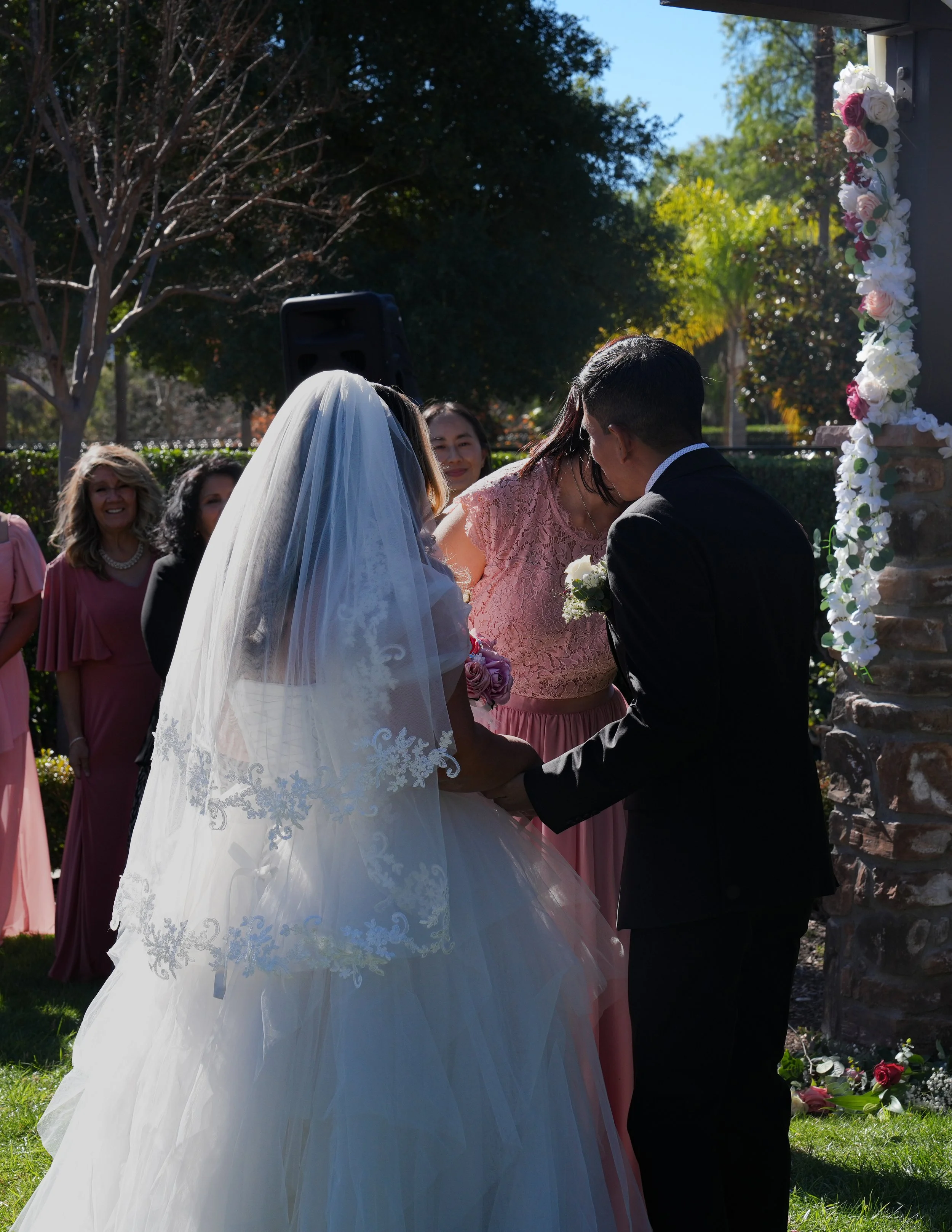 Wedding ceremony with bride in white gown and veil and groom in black suit holding hands, surrounded by women in pink dresses and guests outside, decorated with flowers.
