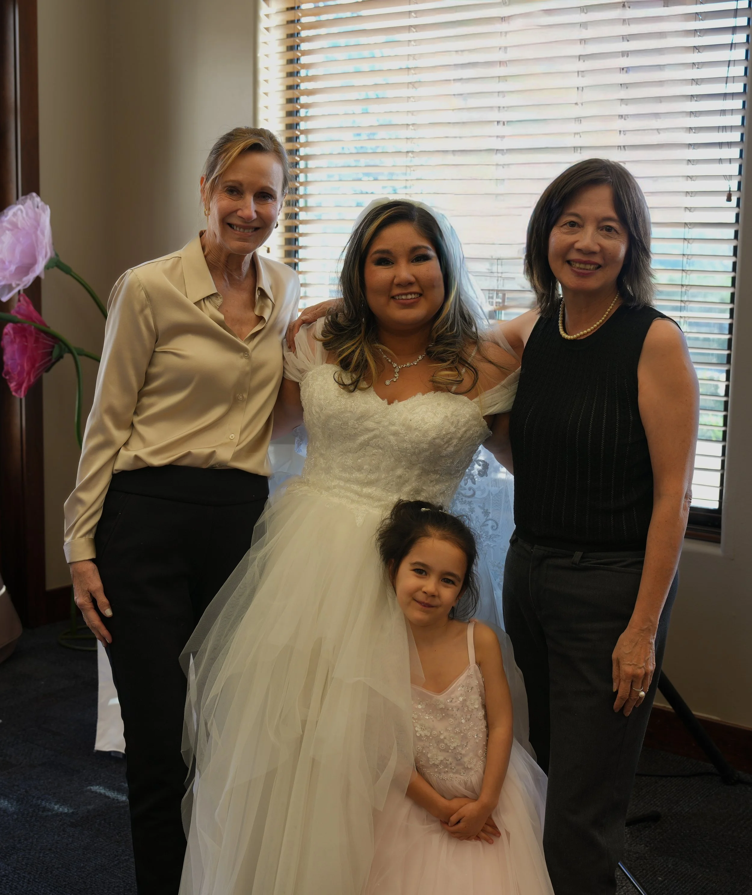 Four women, including a bride in a wedding dress and a young girl in a dress, smiling and standing together indoors near a window with blinds.