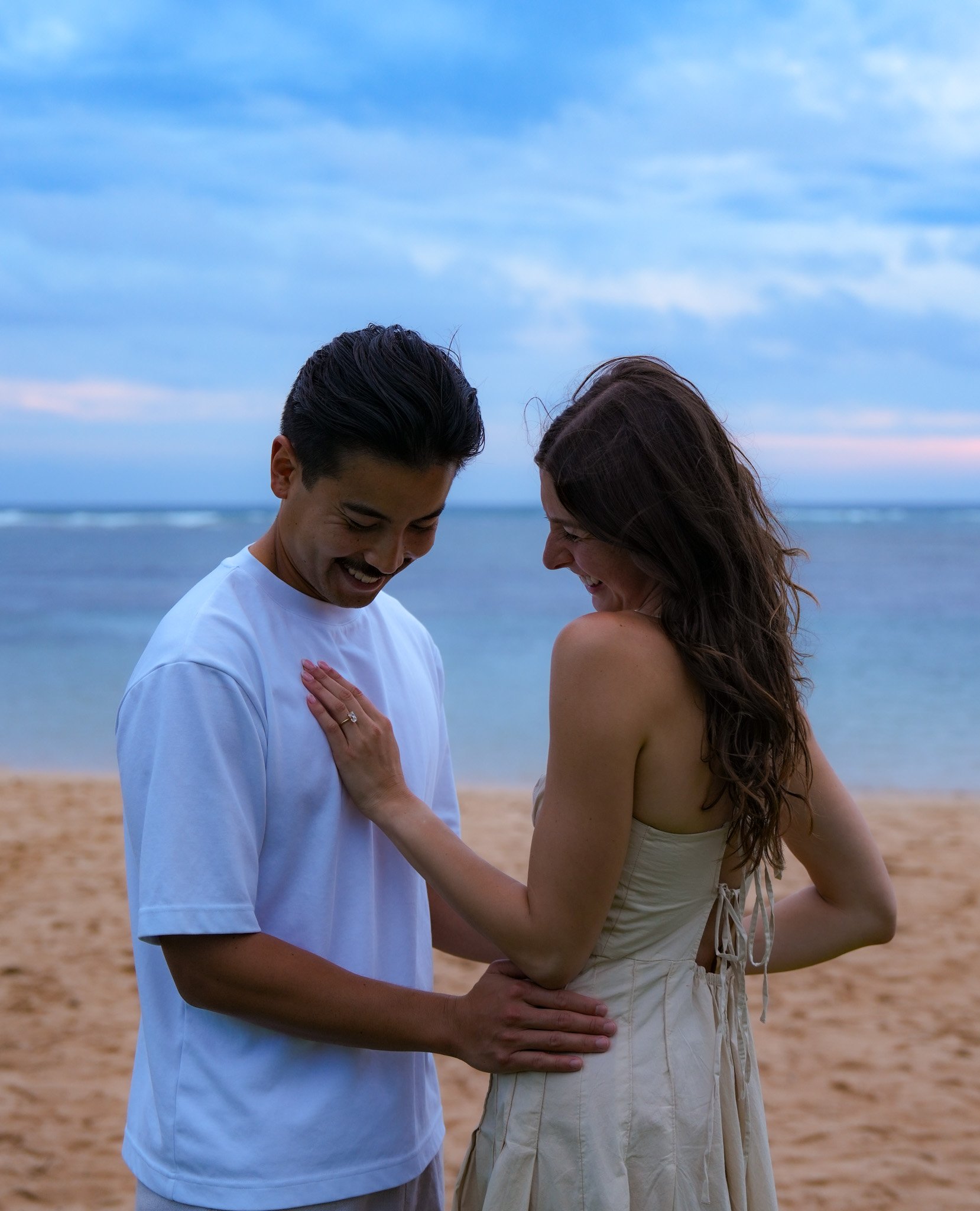 A couple standing on the beach, smiling and laughing together, with the ocean and sky in the background.