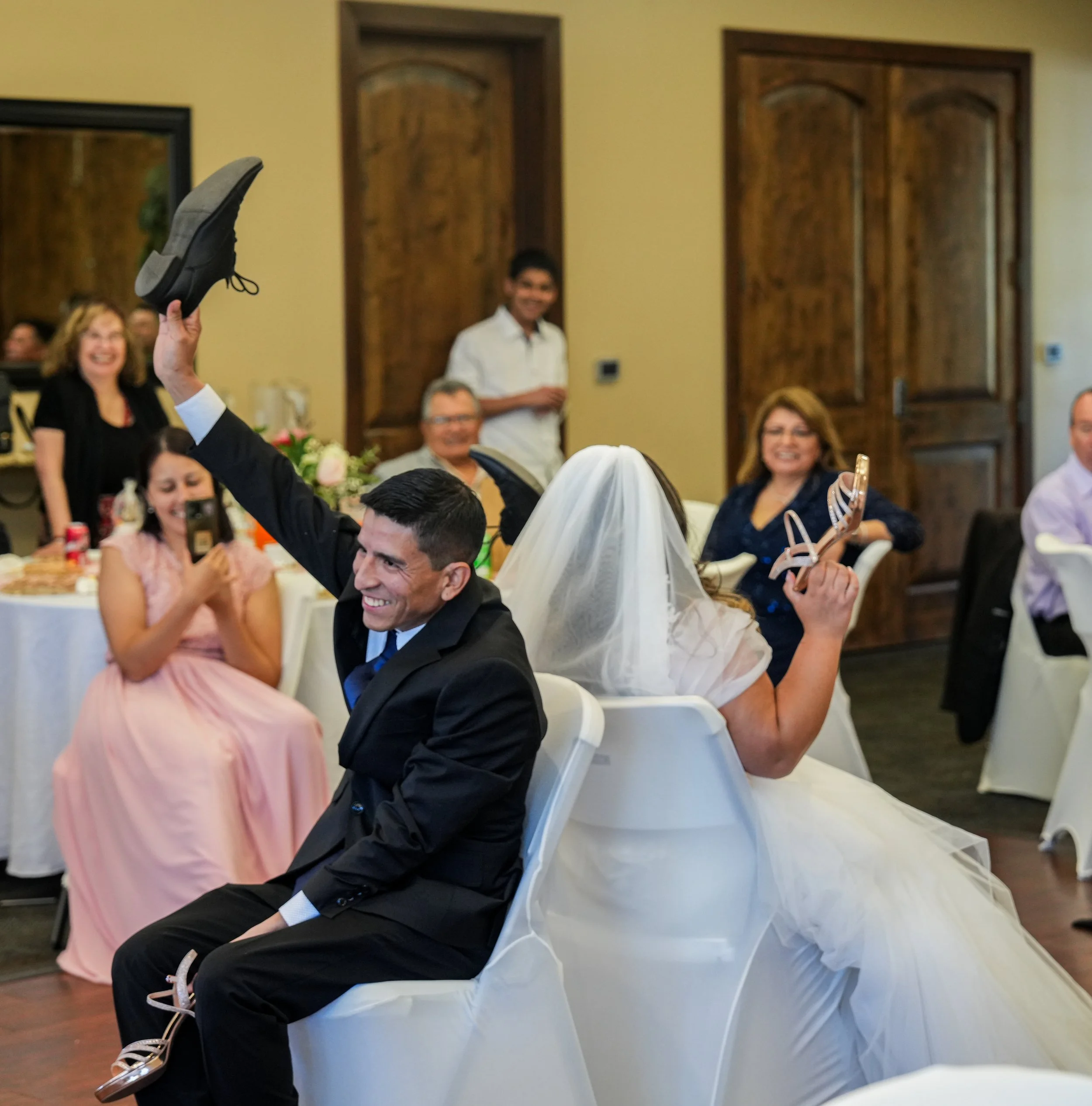 A wedding reception with a bride and groom sitting back-to-back and playing a game, each holding a shoe. The bride is wearing a white dress and veil, and the groom is in a black suit. Guests are smiling and enjoying the moment in the background.