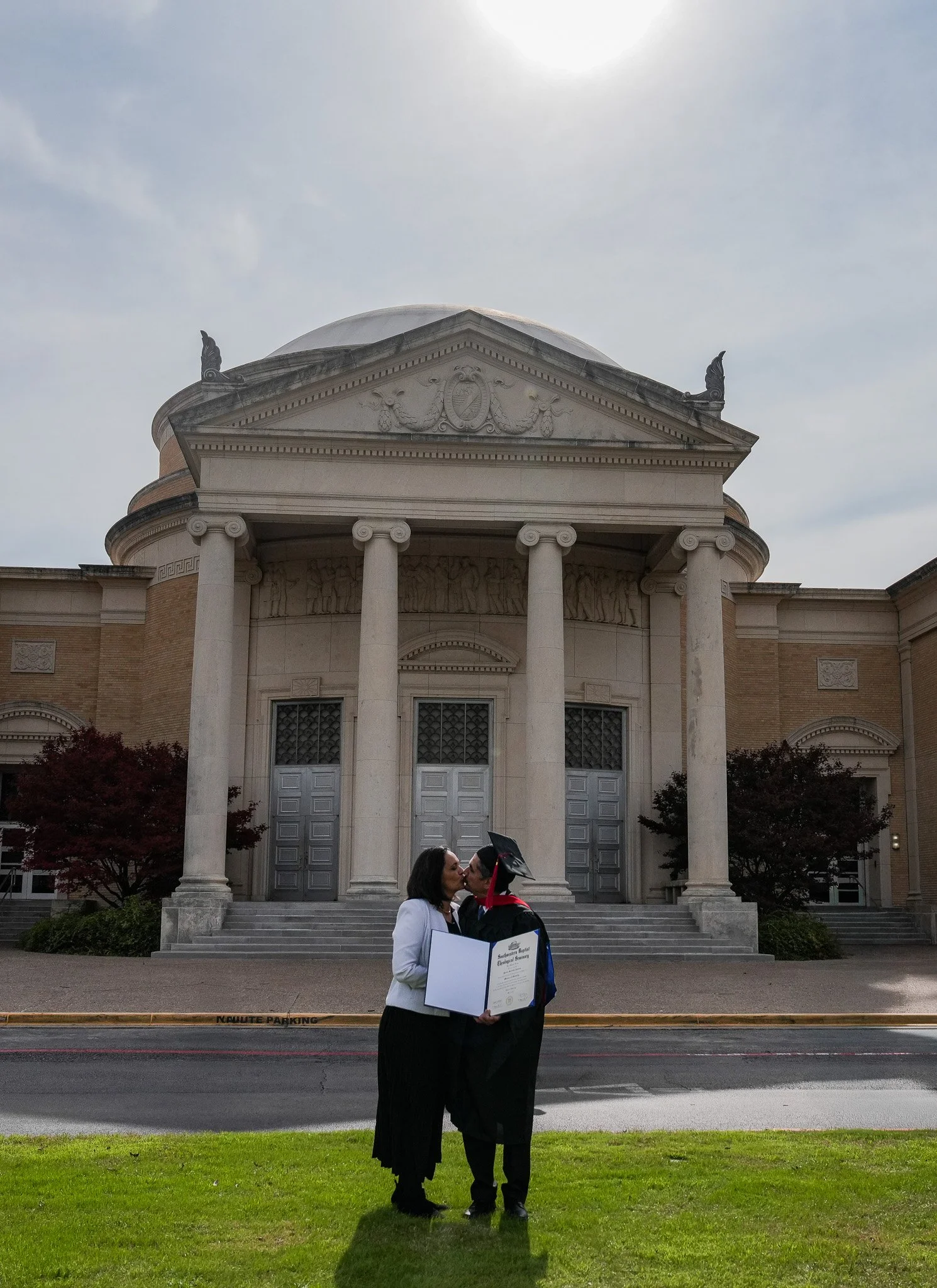 A graduate in black cap and gown kissing a woman holding a diploma in front of a large, classical-style building with columns.