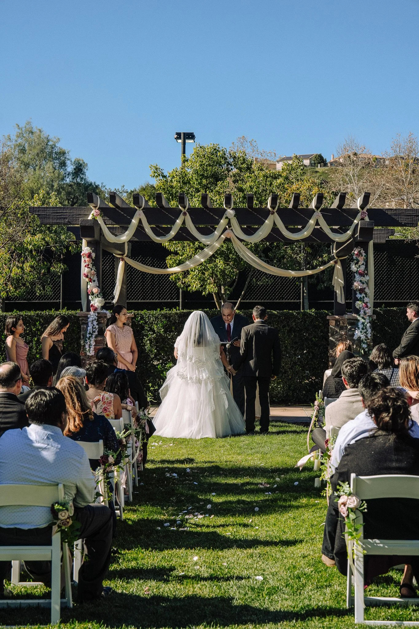 Outdoor wedding ceremony with a bride and groom standing before an officiant, under a decorated wooden arch, with guests seated on either side, in a garden setting.