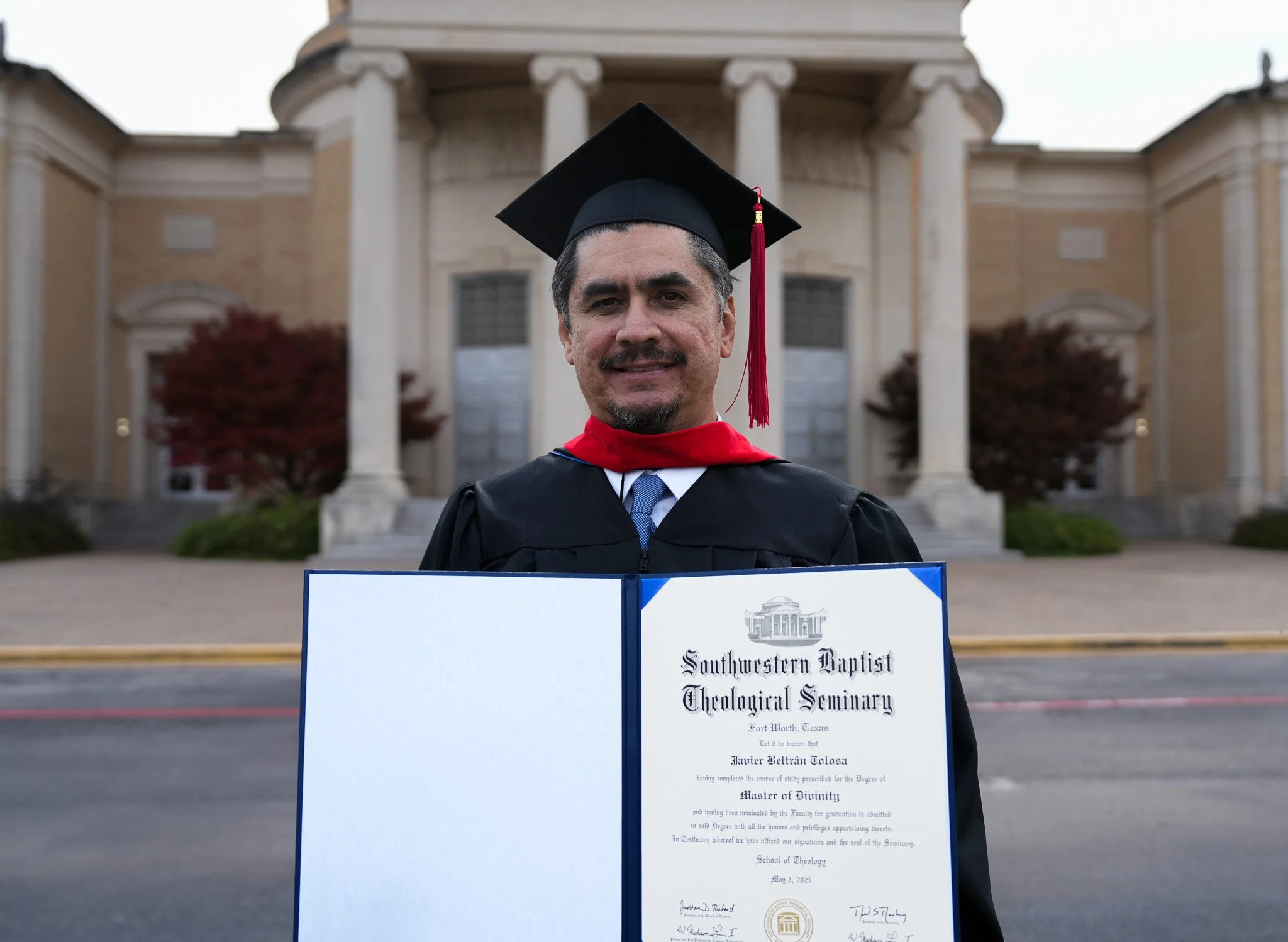 A man in graduation cap and gown holding a diploma standing in front of a church-like building.