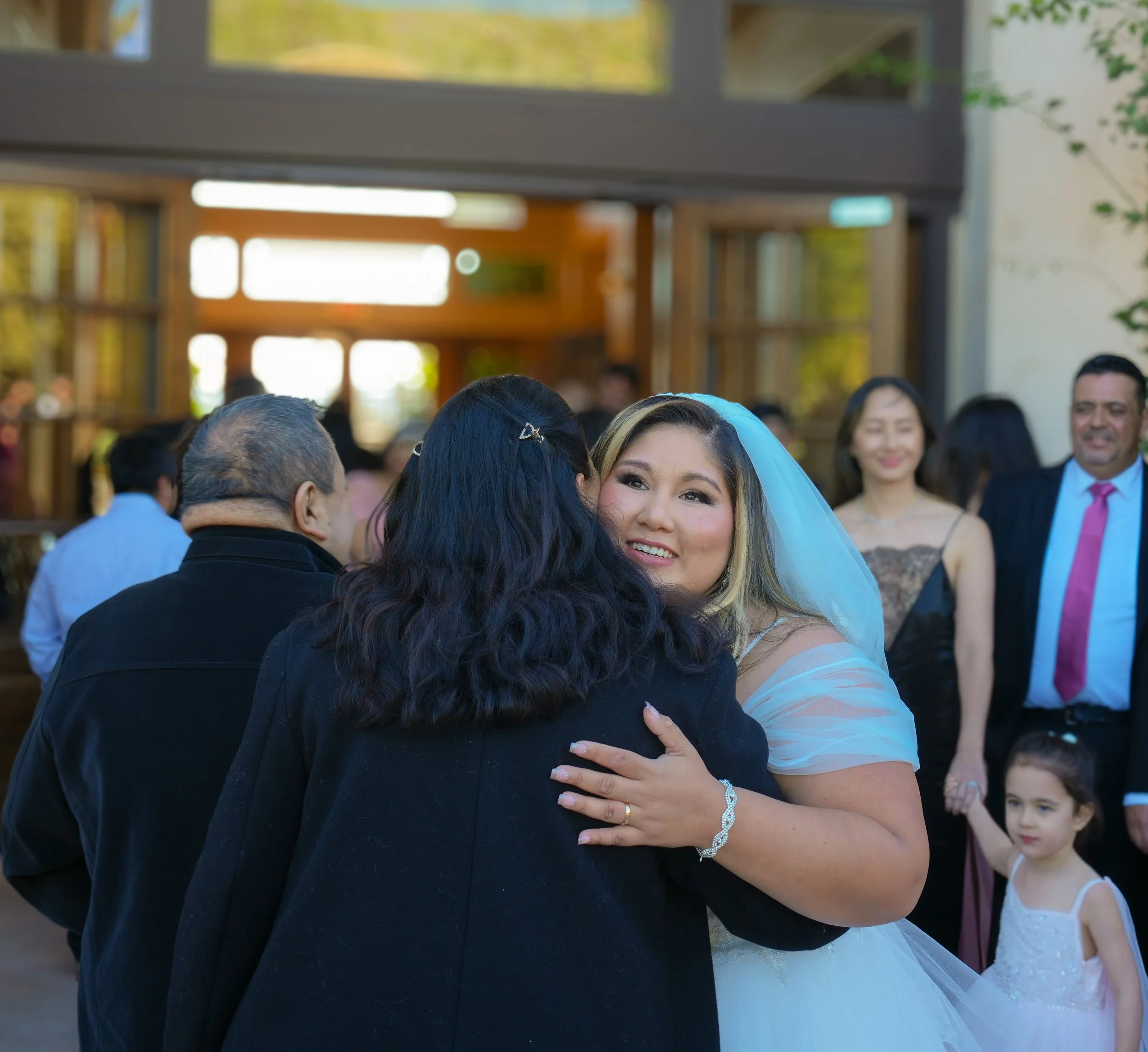 A bride hugging a woman at a wedding reception with guests in the background.