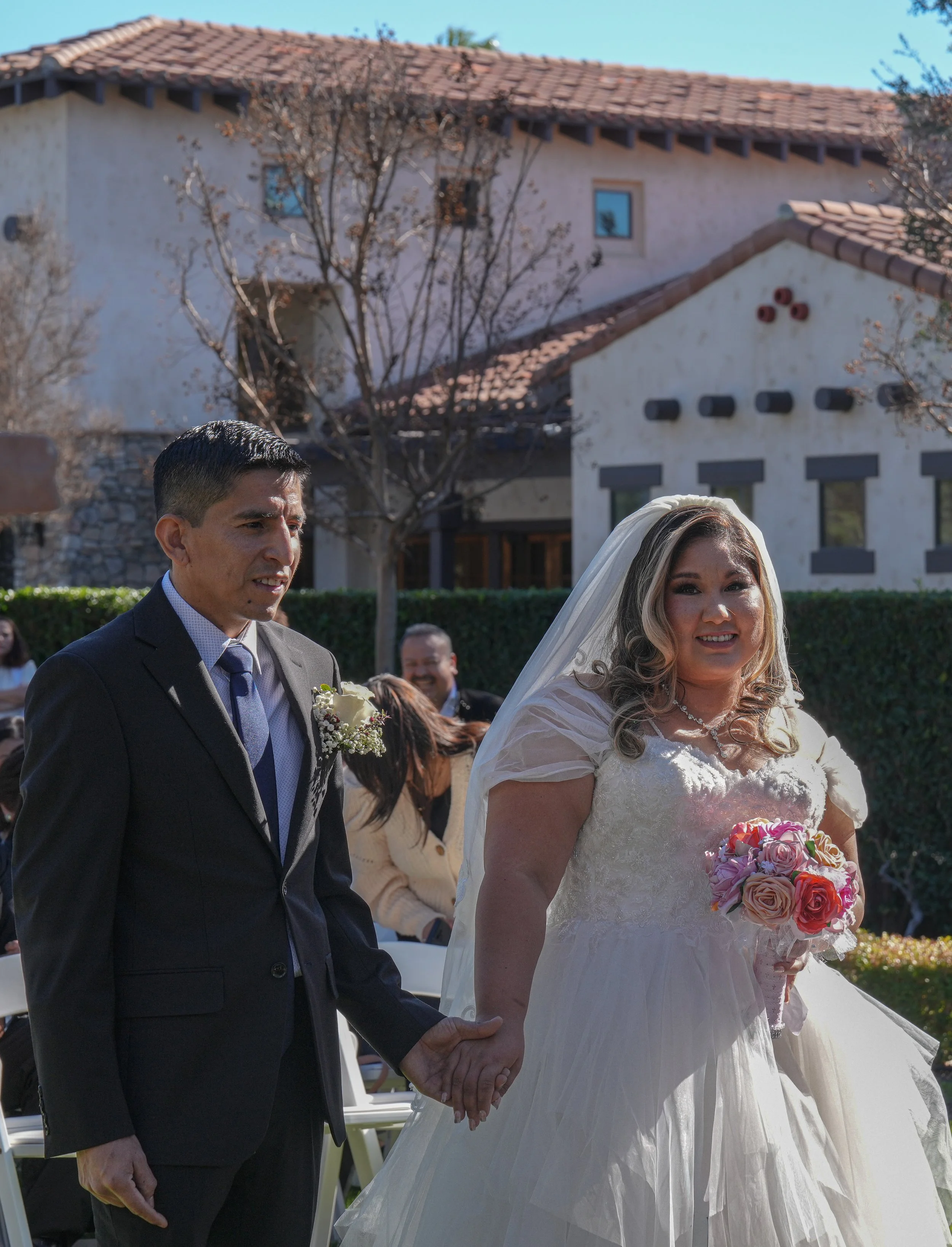 A bride and groom holding hands during their wedding ceremony outdoors, with guests in the background and a house with a tiled roof and trees behind them.