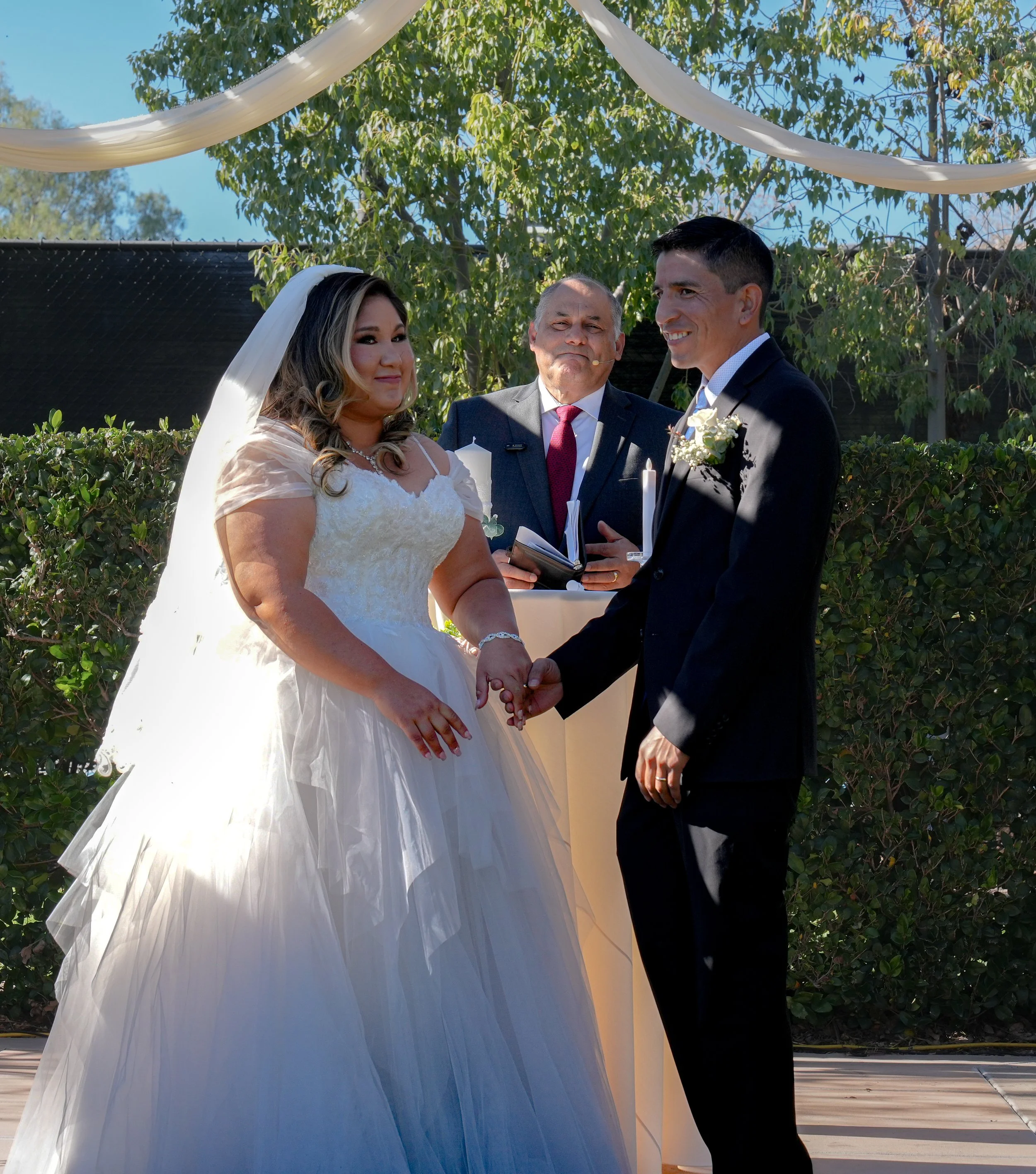A couple getting married outdoors, holding hands and smiling at each other, with an officiant standing behind them, all under a white fabric canopy and surrounded by greenery.