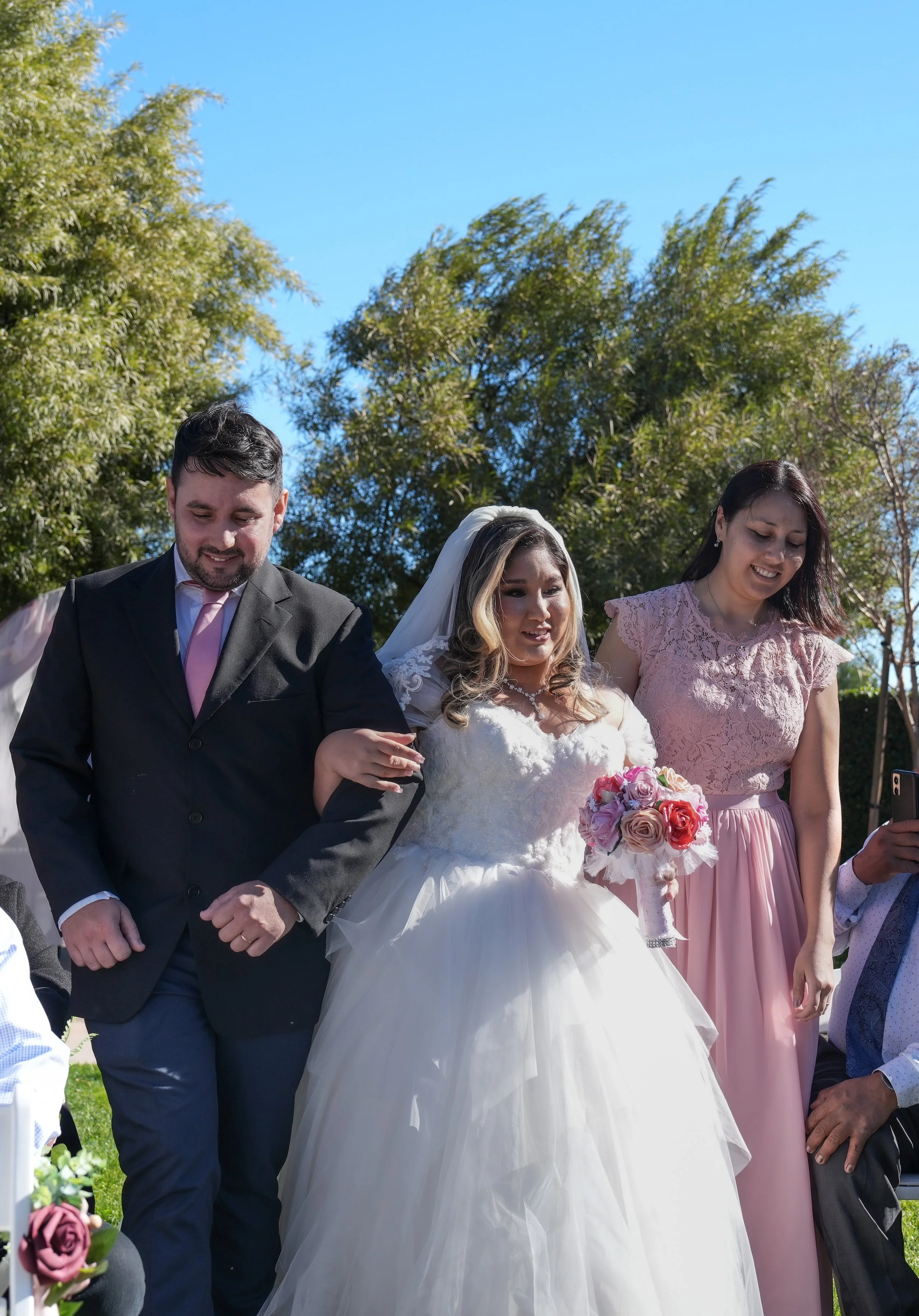 A bride in a white wedding gown holding a bouquet of pink and purple flowers, walking outdoors with a man in a black suit and a woman in a pink dress, surrounded by greenery and blue sky.