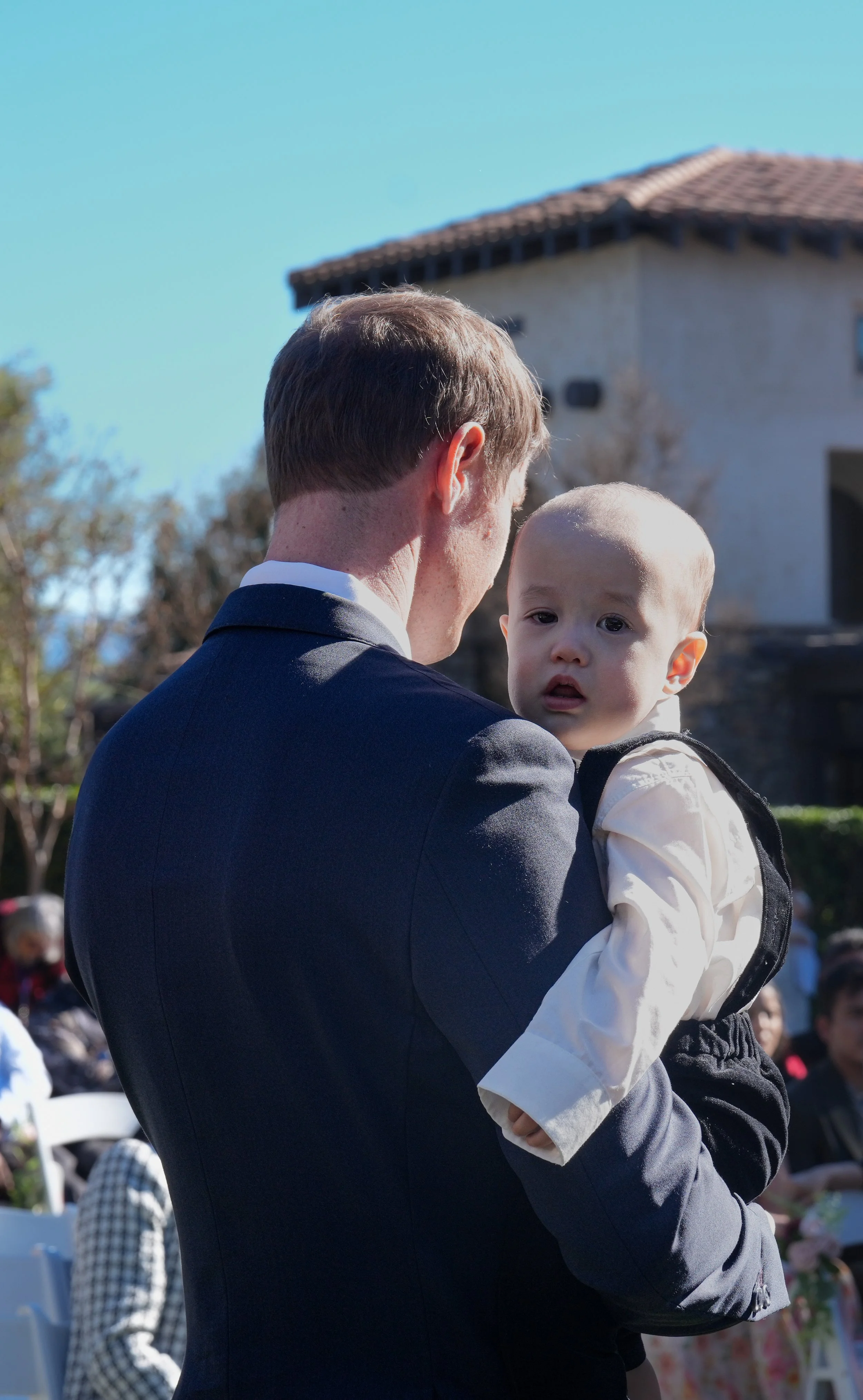 A man in a suit holding a young boy in a white shirt outdoors on a sunny day.