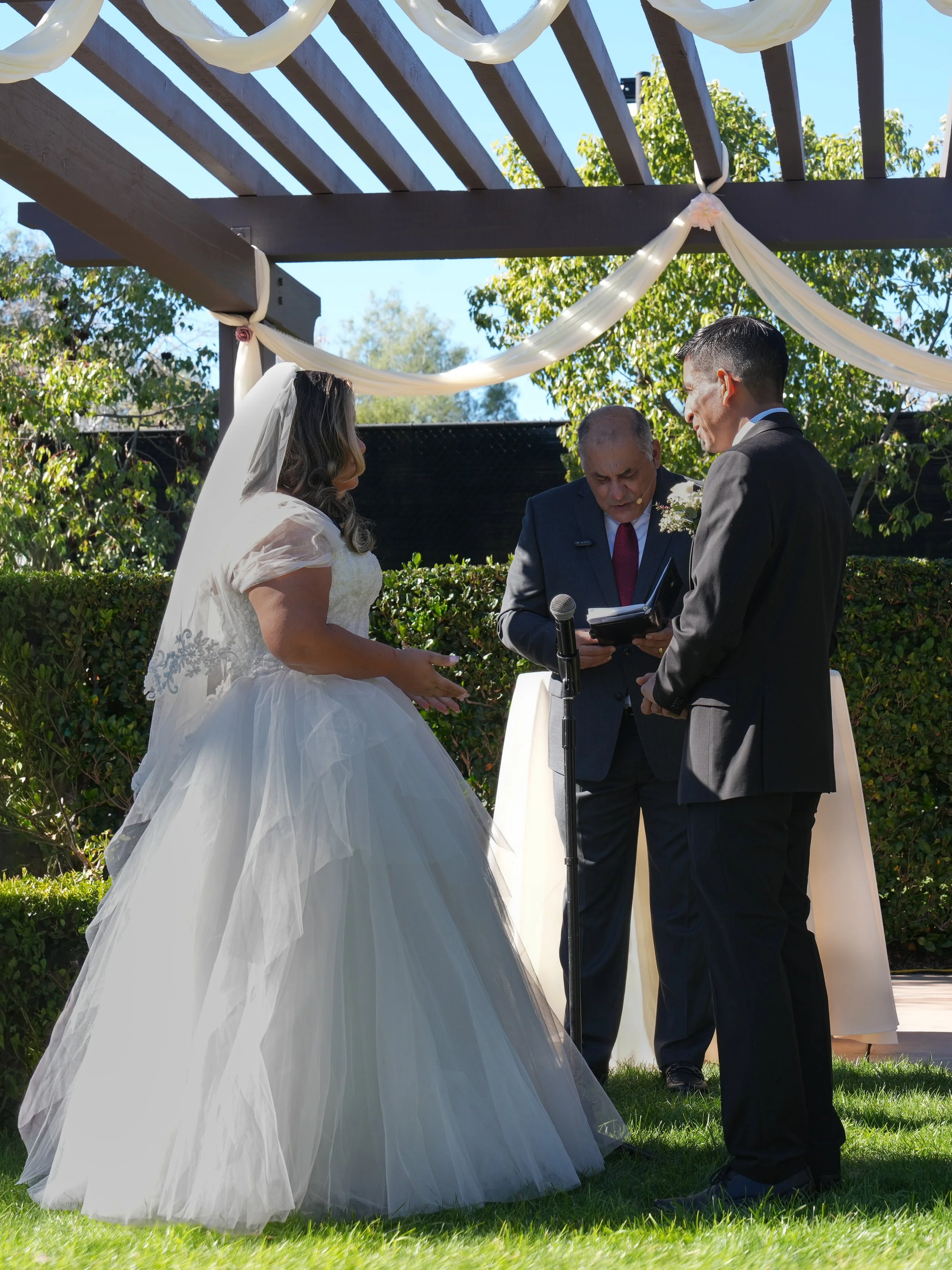 A wedding ceremony taking place outdoors under a wooden pergola decorated with white fabric. A bride in a white wedding gown and veil, and a groom in a dark suit, are exchanging vows in front of an officiant dressed in a dark suit. The ceremony is be