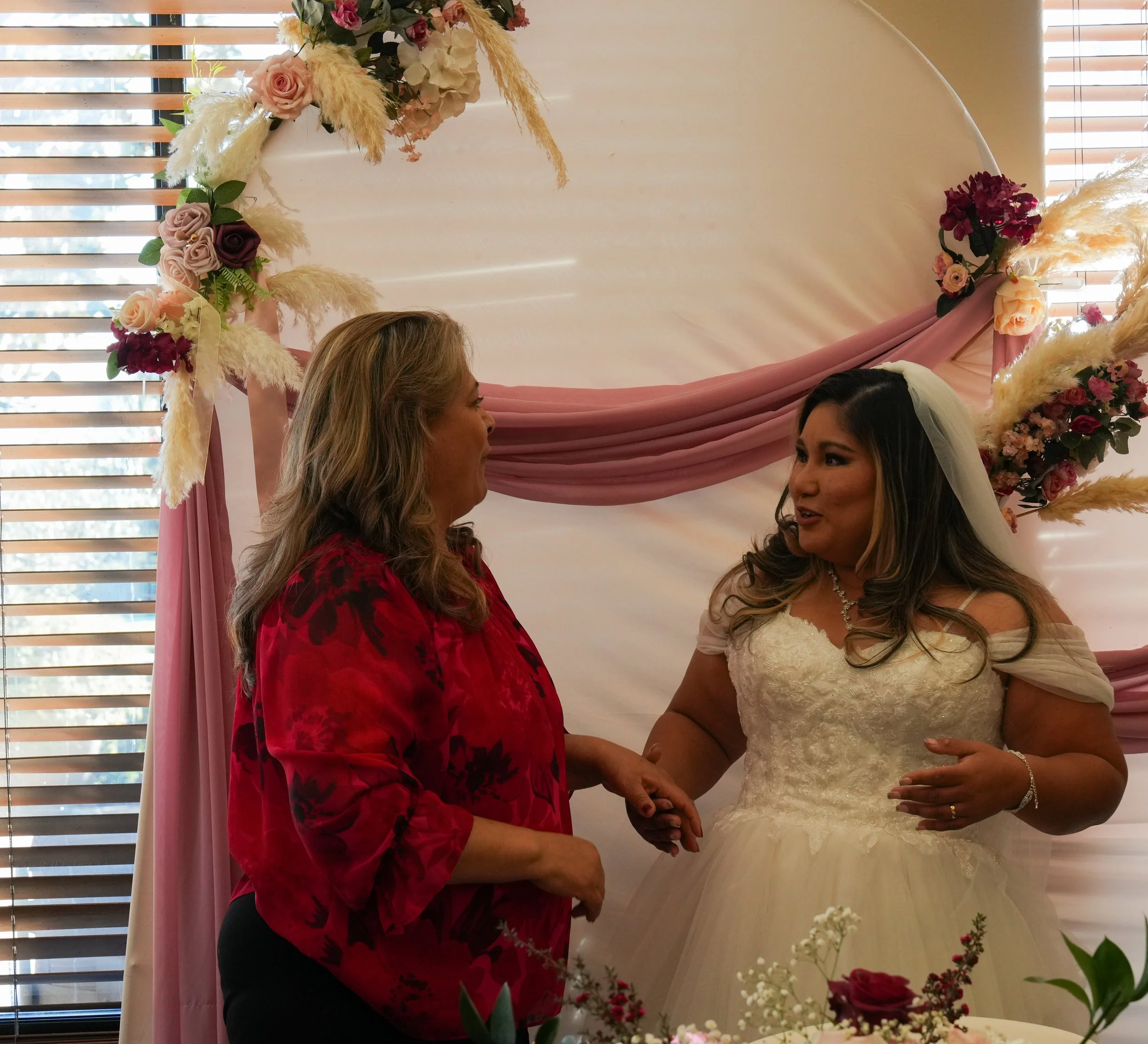 A woman in a red shirt talking to a bride in a white wedding dress, with floral decorations and pink drapery in the background.