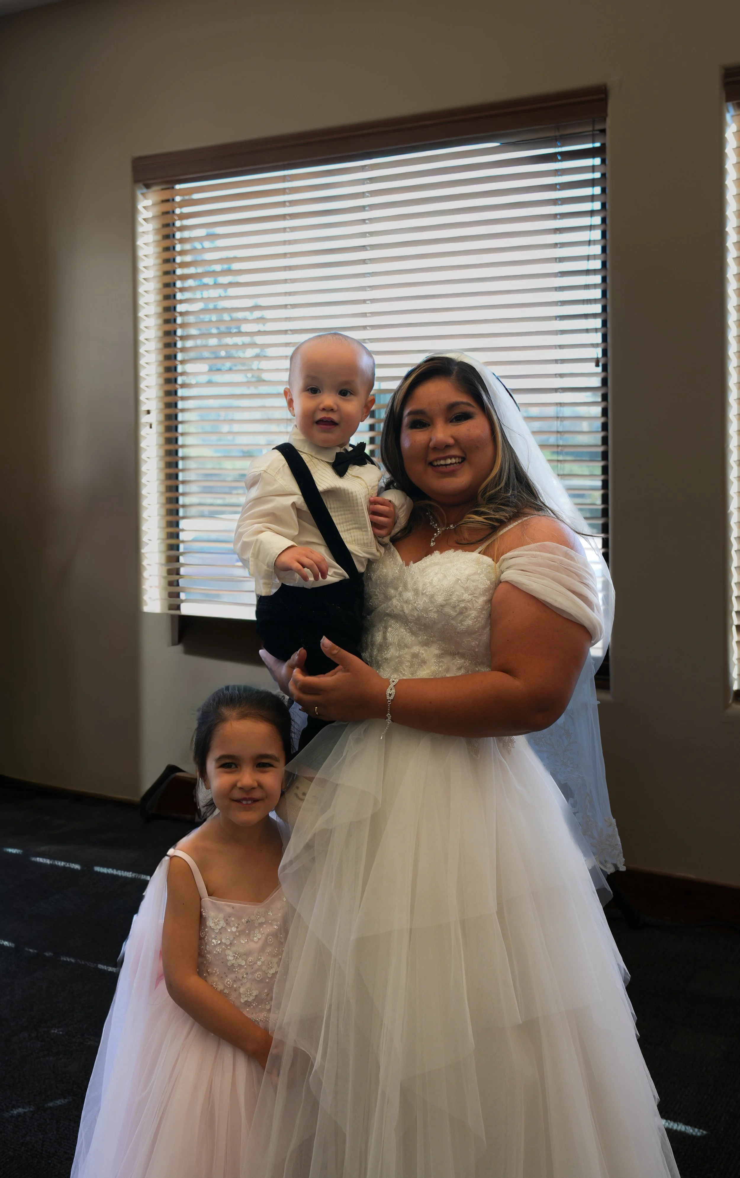 A bride with a baby boy and a young girl posing indoors with window blinds in the background.