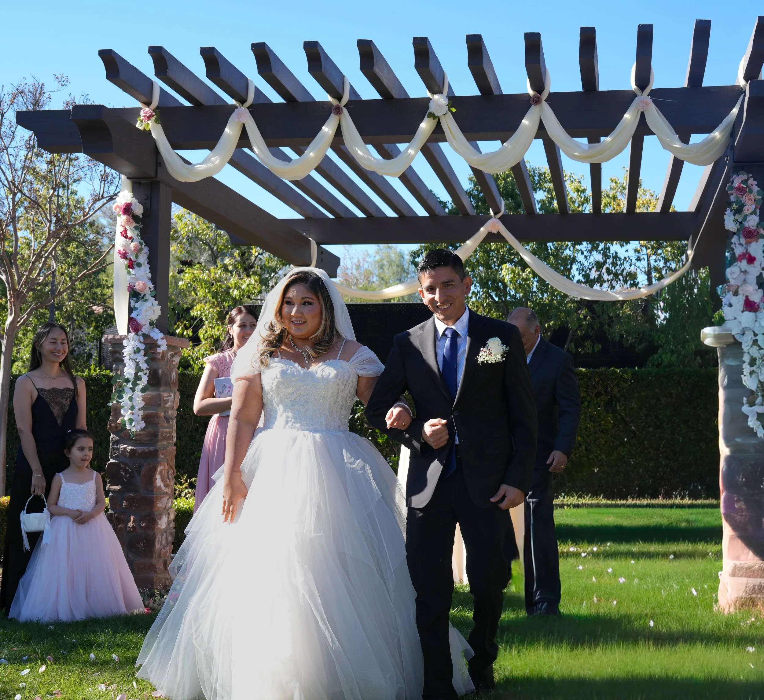Bride and groom walking arm in arm under a decorated outdoor wedding arch with wedding guests in the background.