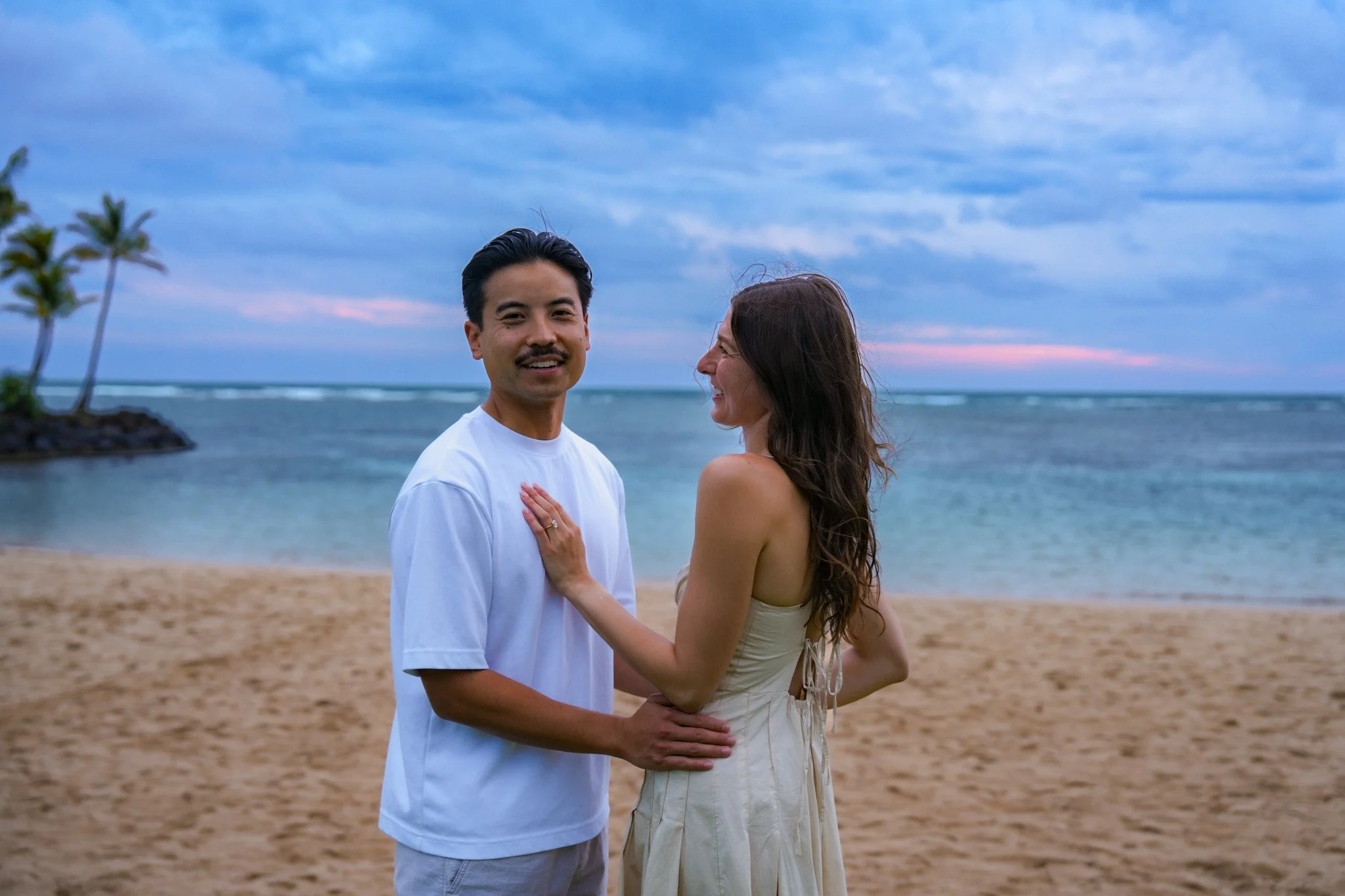 A couple standing on a beach at sunset, with palm trees in the background, smiling and looking at each other while holding each other's waists.