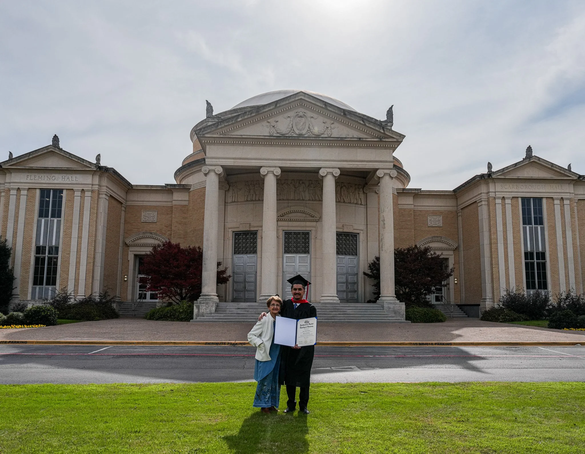 A graduate in a gown and cap holding a diploma, standing next to an older woman outside a classical style building with columns, labeled Fleming Hall and Scarborough Hall, during daytime with a gray sky.