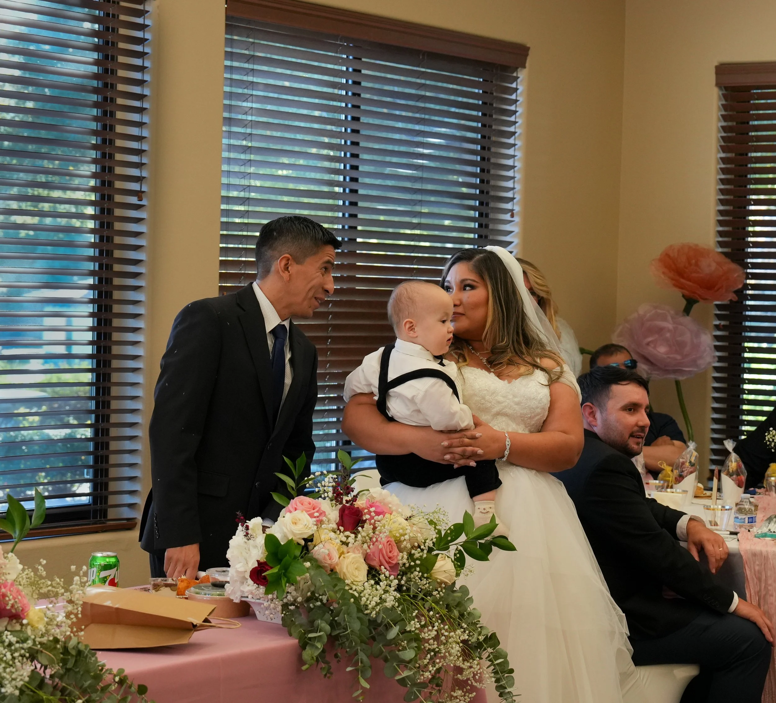 A bride holding a young boy at a wedding reception, with a man talking to her, seated guests, and floral decorations in the background.