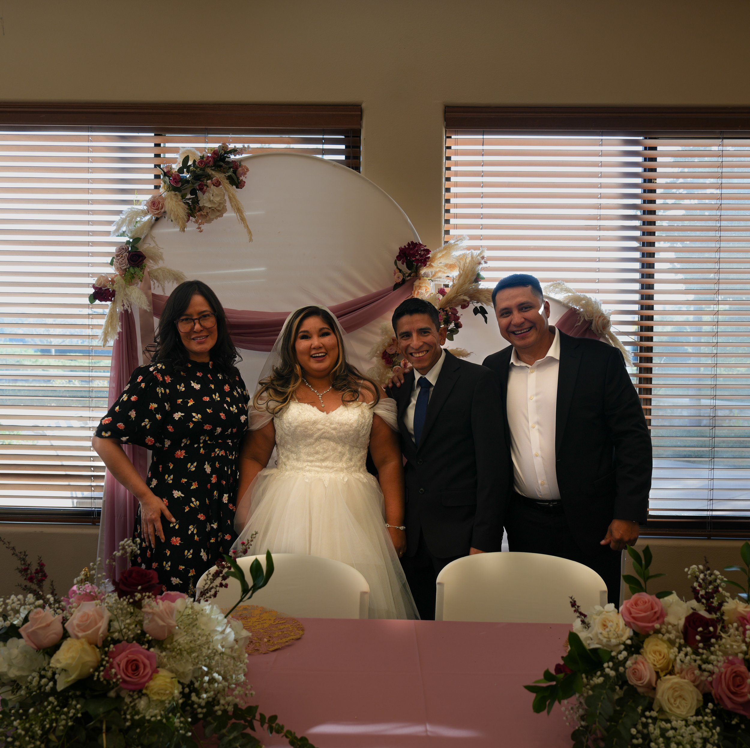 Four people, including a bride in a white wedding dress and groom in a black suit, smiling and standing together in front of a decorated backdrop at a wedding celebration, with floral arrangements and decorated arch behind them.