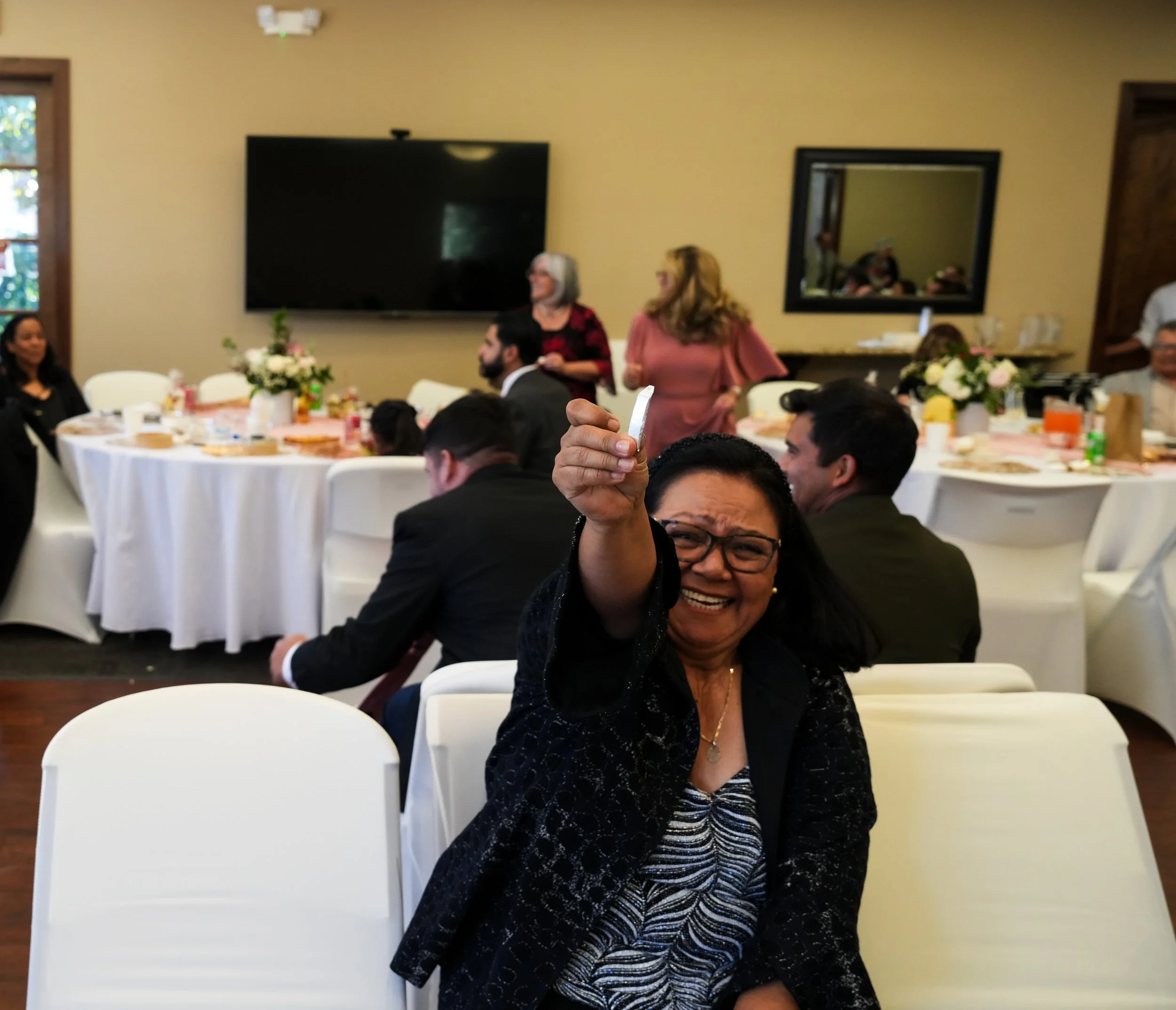 A woman smiling and laughing, pointing toward the camera, at a celebratory gathering with tables set for a meal and other people engaging in conversation in the background.