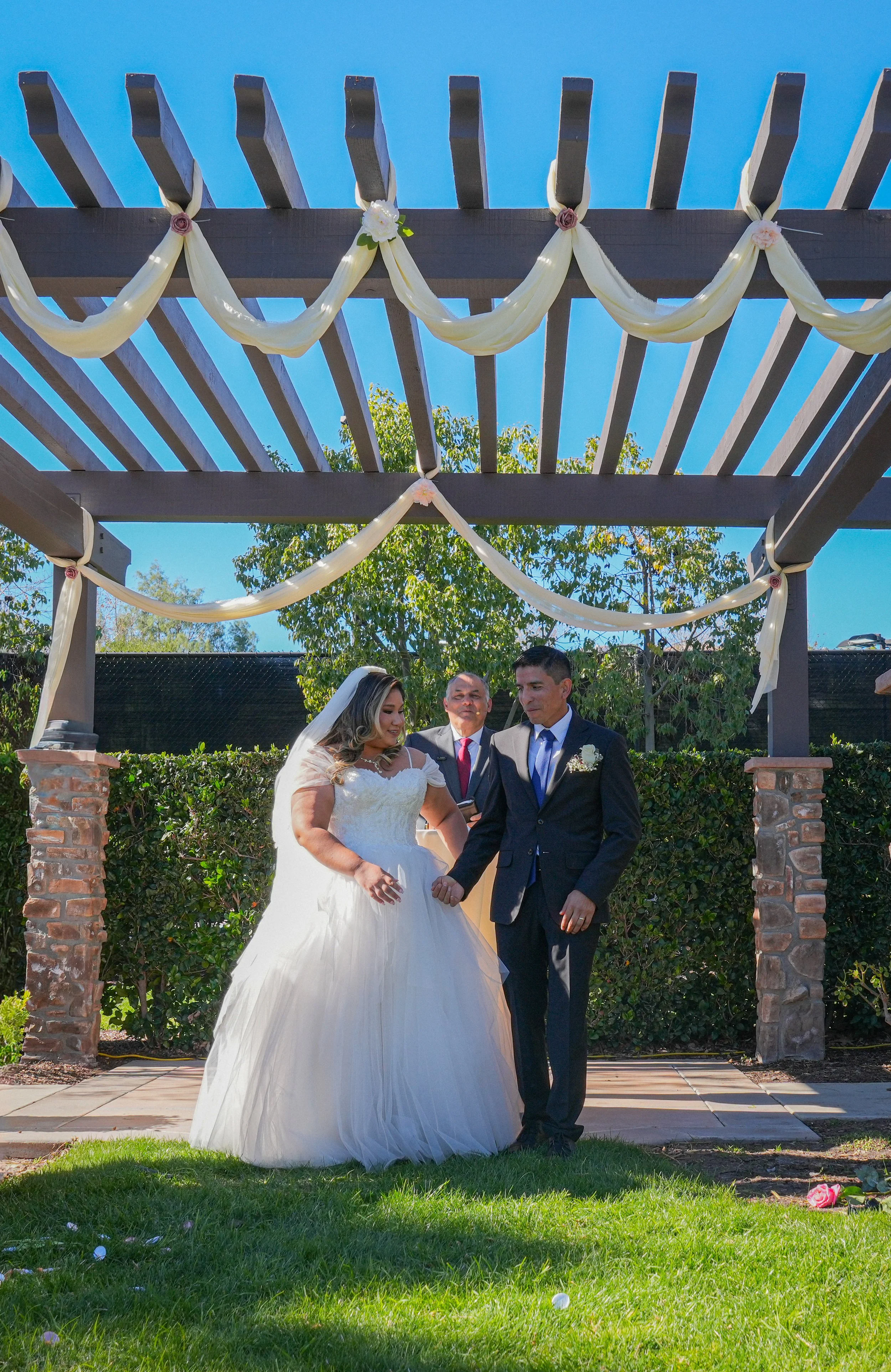 A bride and groom holding hands during their wedding ceremony outdoors, with a officiant behind them, under a wooden pergola decorated with white fabric and pink flowers, on a sunny day with green trees in the background.