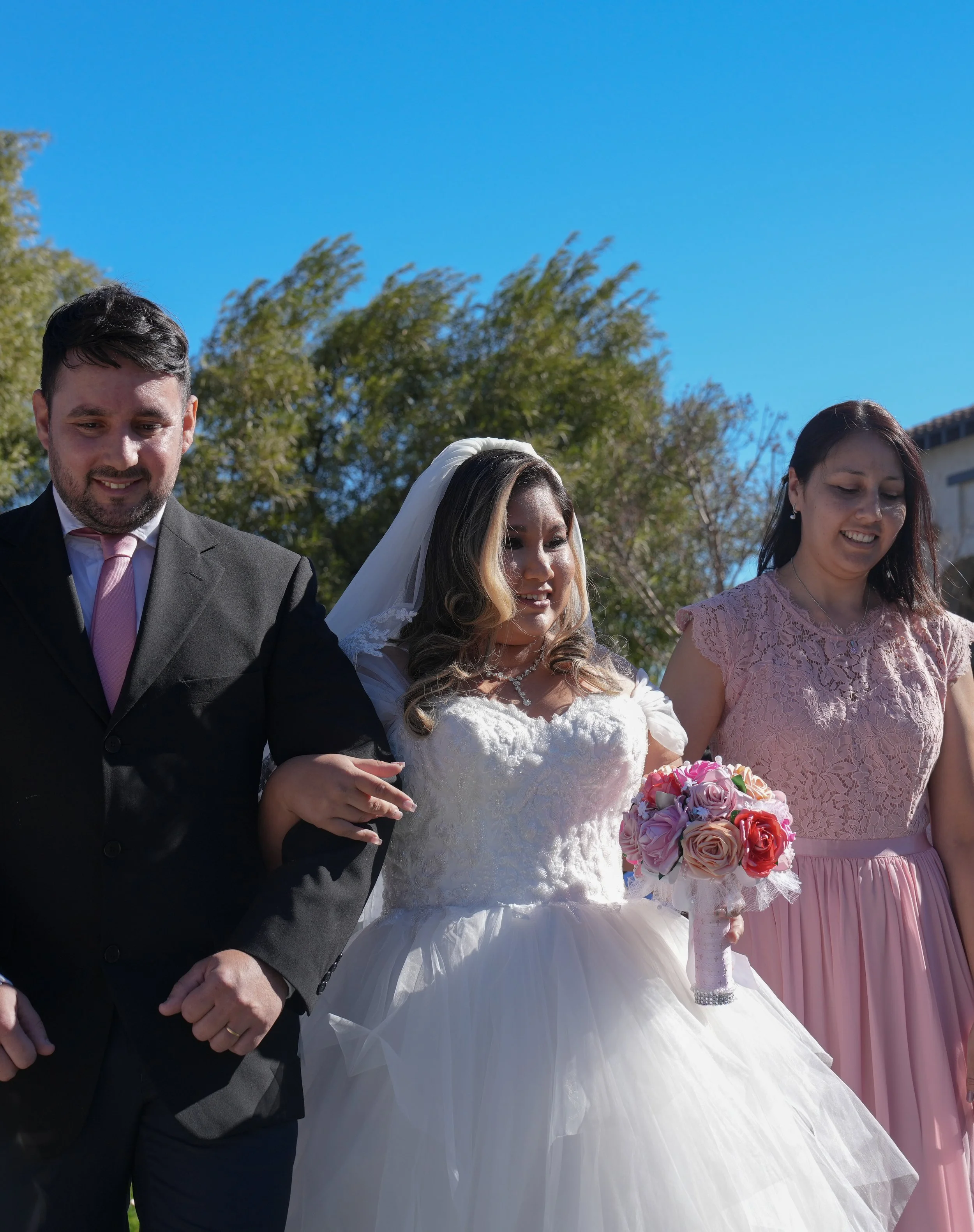 Bride in a white wedding dress holding a pink flower bouquet, flanked by a man in a black suit and woman in a pink dress, outdoors on a sunny day.