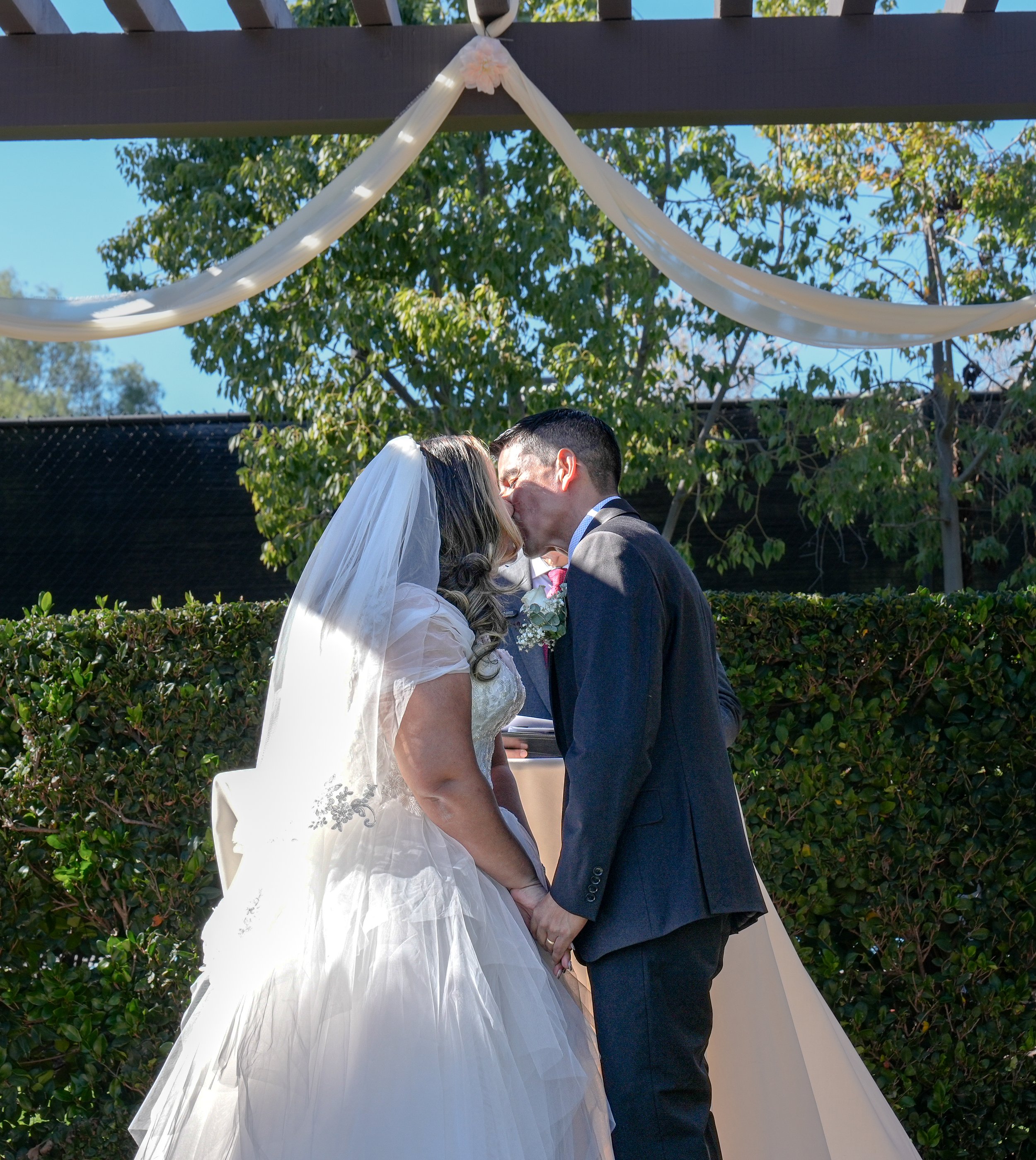 A bride and groom kissing during their wedding ceremony outdoors, with trees and a decorated backdrop in the background.