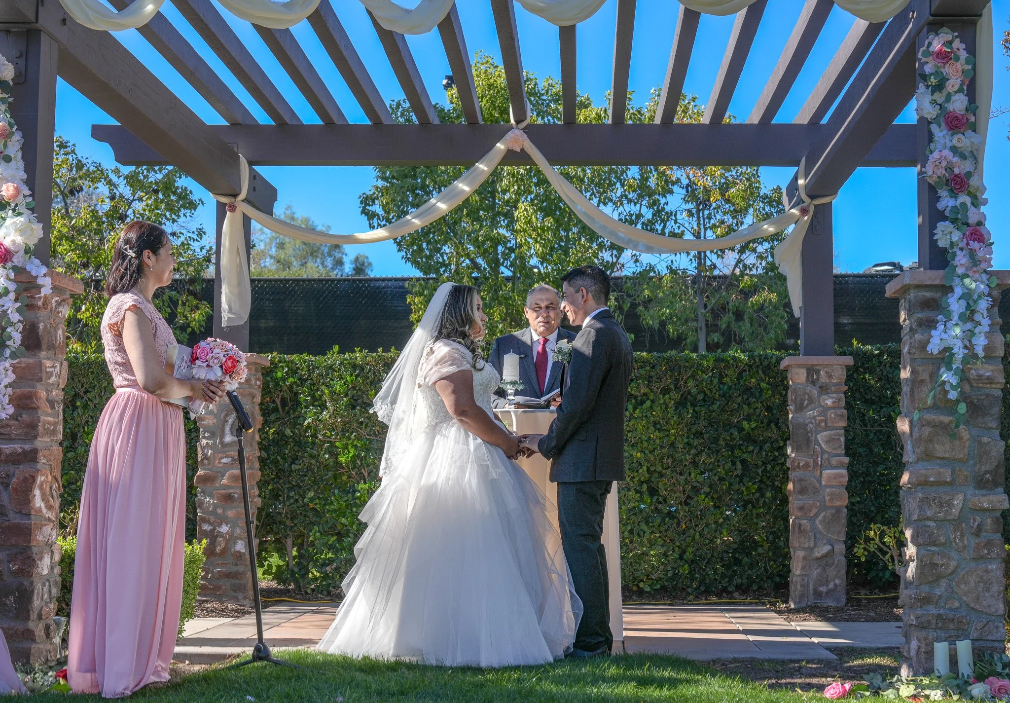 A wedding ceremony outdoors with a bride and groom holding hands, an officiant, and a bridesmaid holding a bouquet, under a wooden arch decorated with flowers and fabric, in a garden setting with trees and bright blue sky.