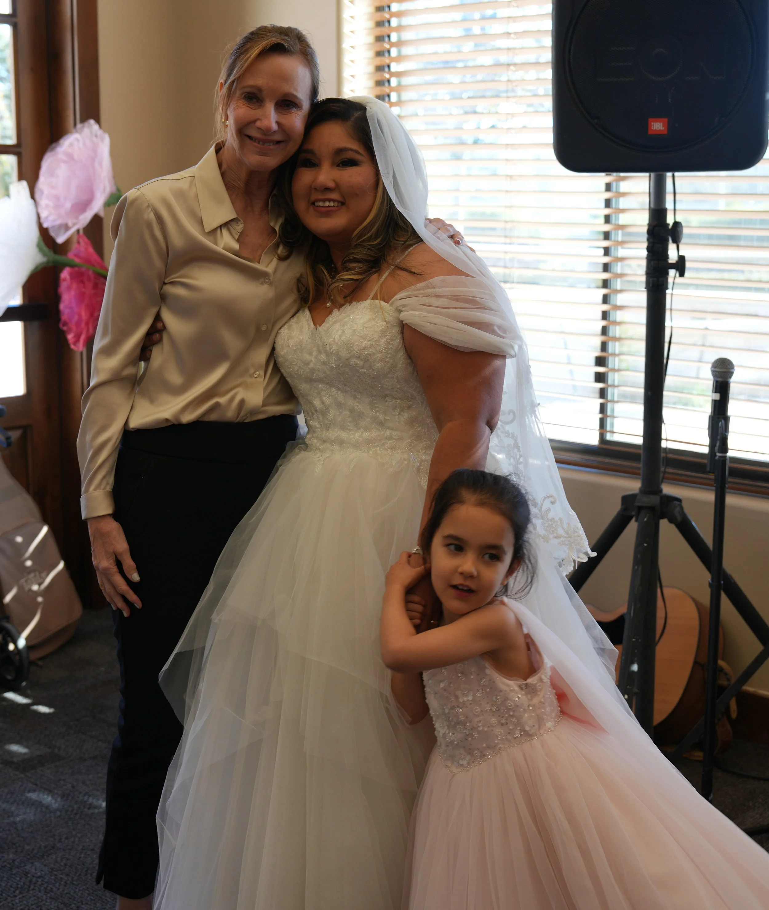 A bride in a white wedding gown and veil, with a woman in a beige blouse and black pants, and a young girl in a pink dress, posing indoors near large windows with blinds. There is a speaker, microphone, and musical guitar visible in the background.