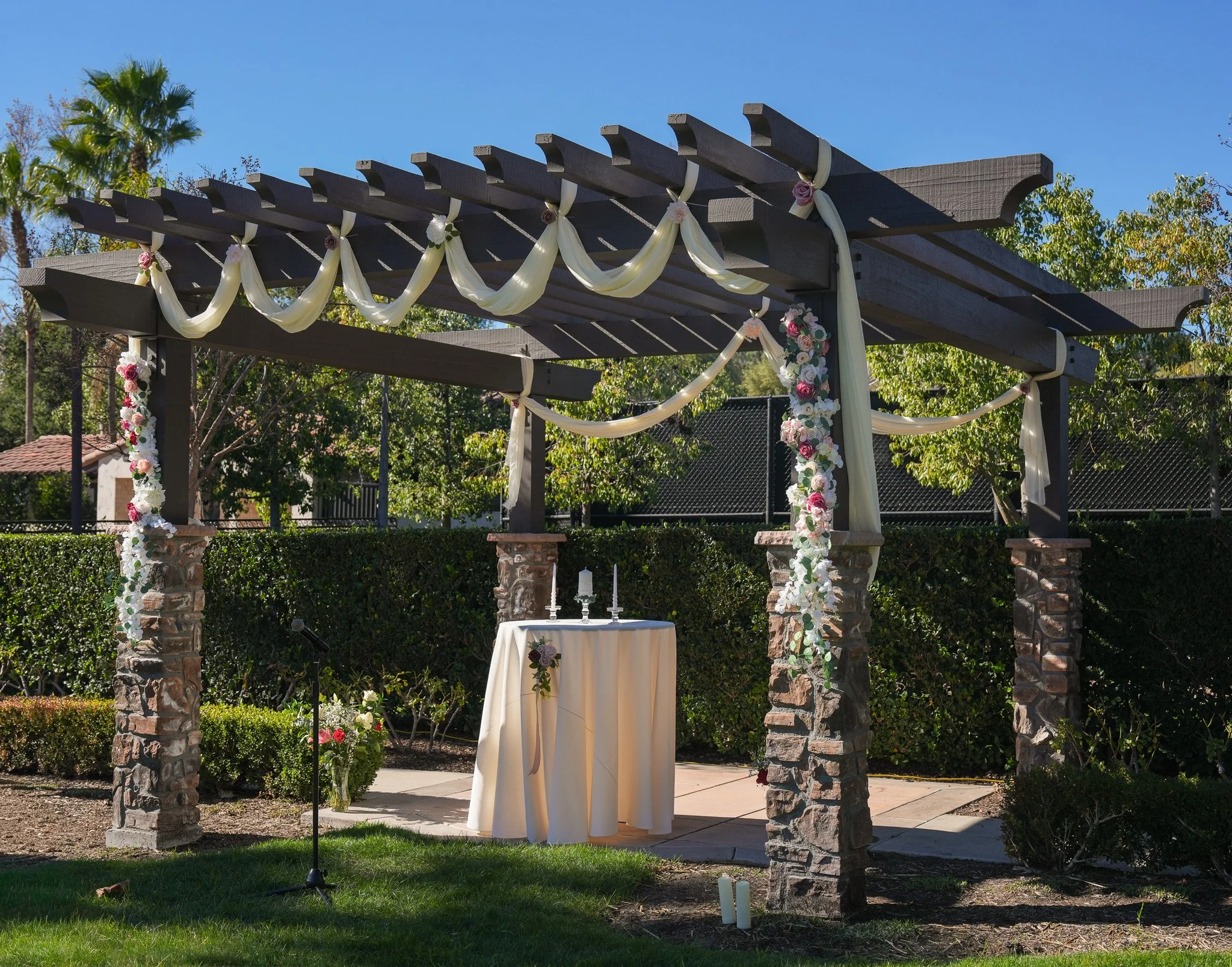 Outdoor wedding setup with a decorated pergola, floral arrangements, a small table with candles, and a microphone stand.