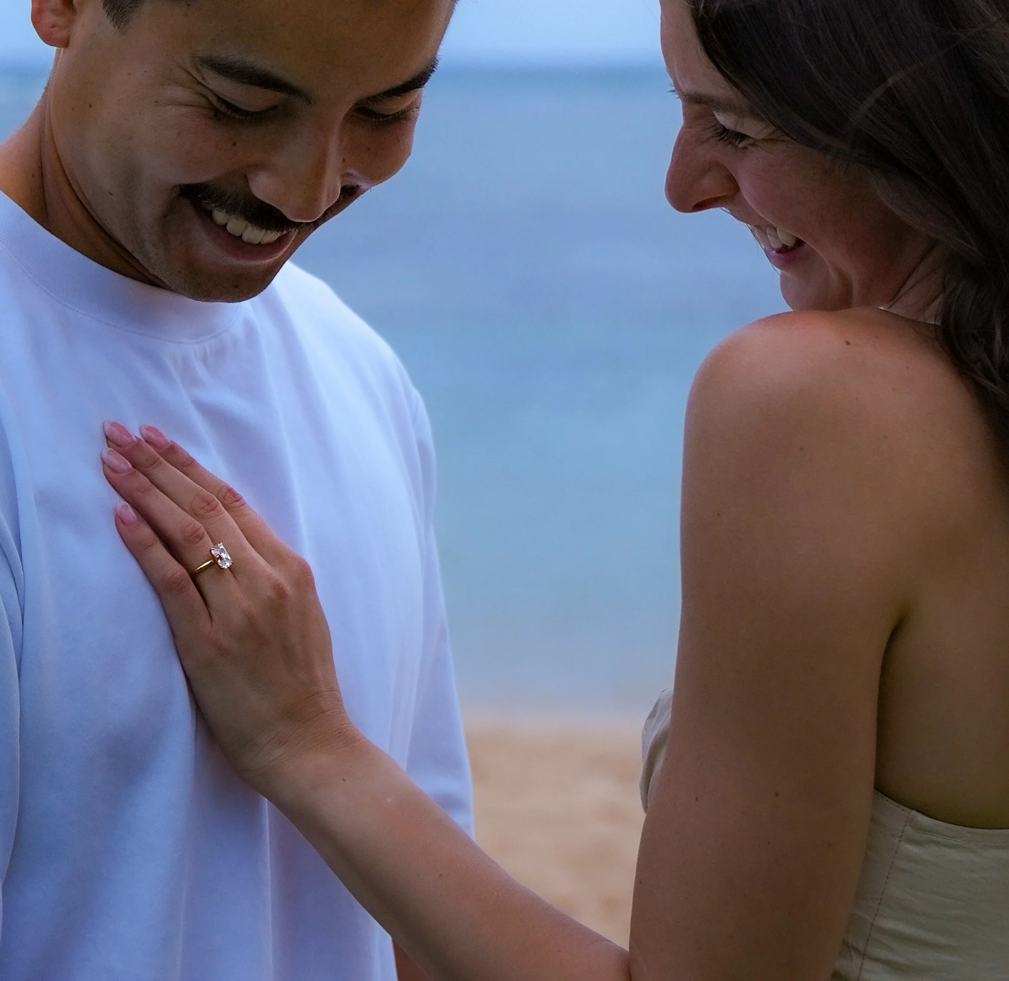 A smiling couple at the beach, with a close-up of the woman's hand showing an engagement ring on her finger, touching the man's chest.