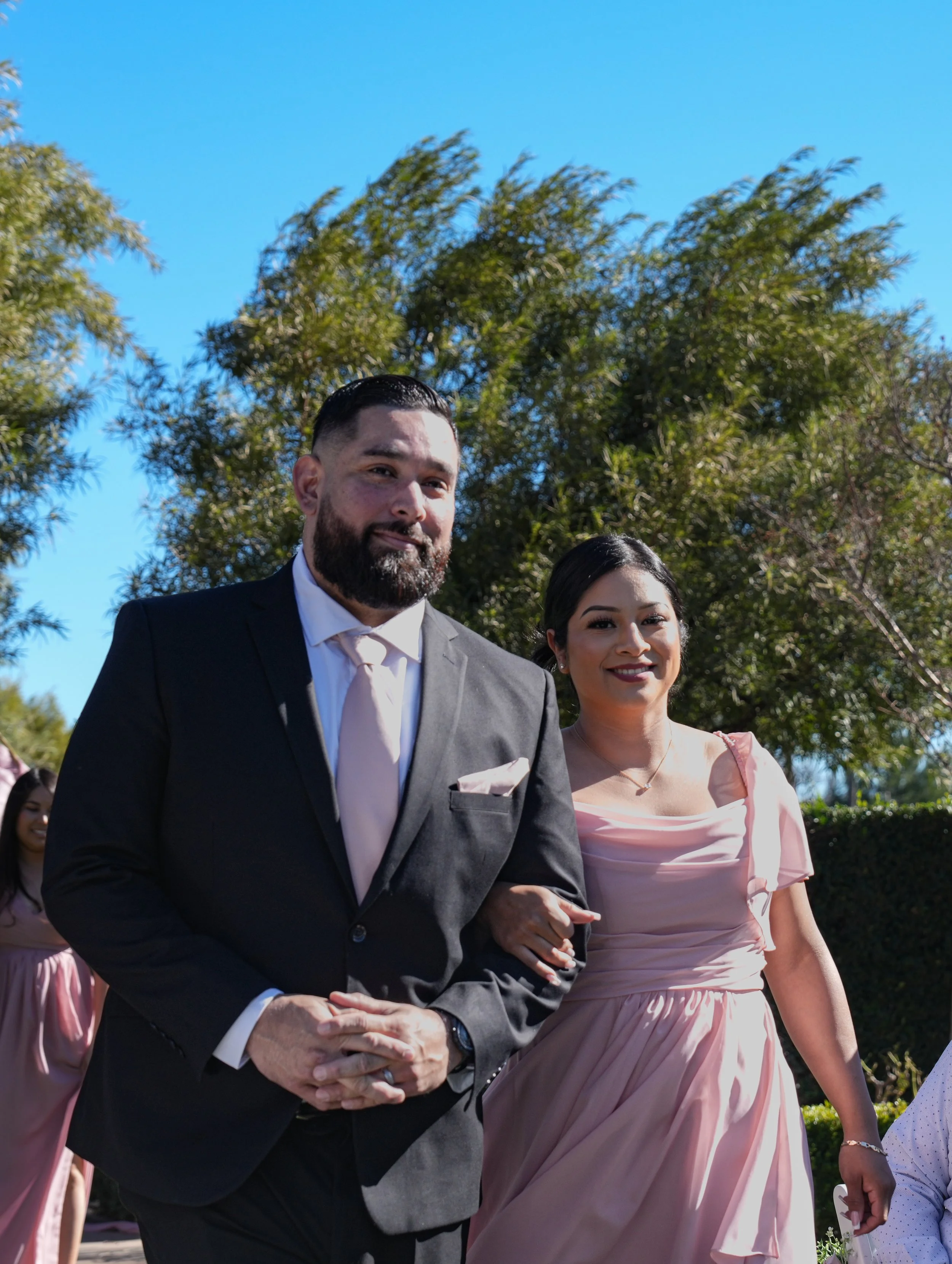 A couple dressed in formal attire, walking outdoors during a sunny day, with a woman in a pink dress visible in the background and green trees and clear blue sky overhead.