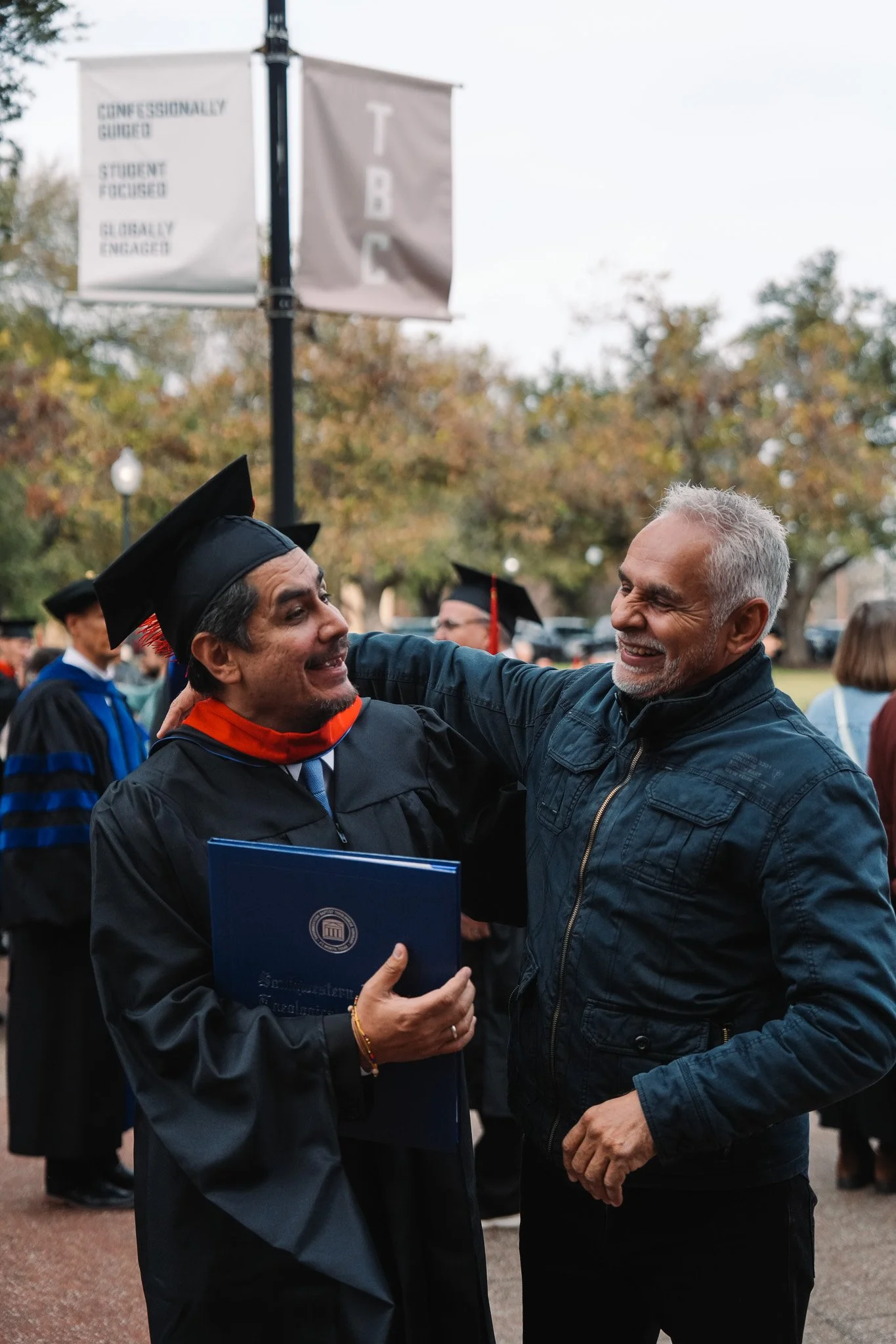 A joyful graduate in a cap and gown celebrating with an older man during a graduation ceremony outdoors, with other graduates in the background and colorful trees.